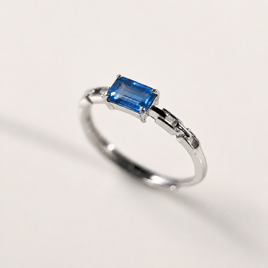 A silver-toned ring featuring a rectangular blue gemstone in a prong setting, photographed on a clean off-white background.
