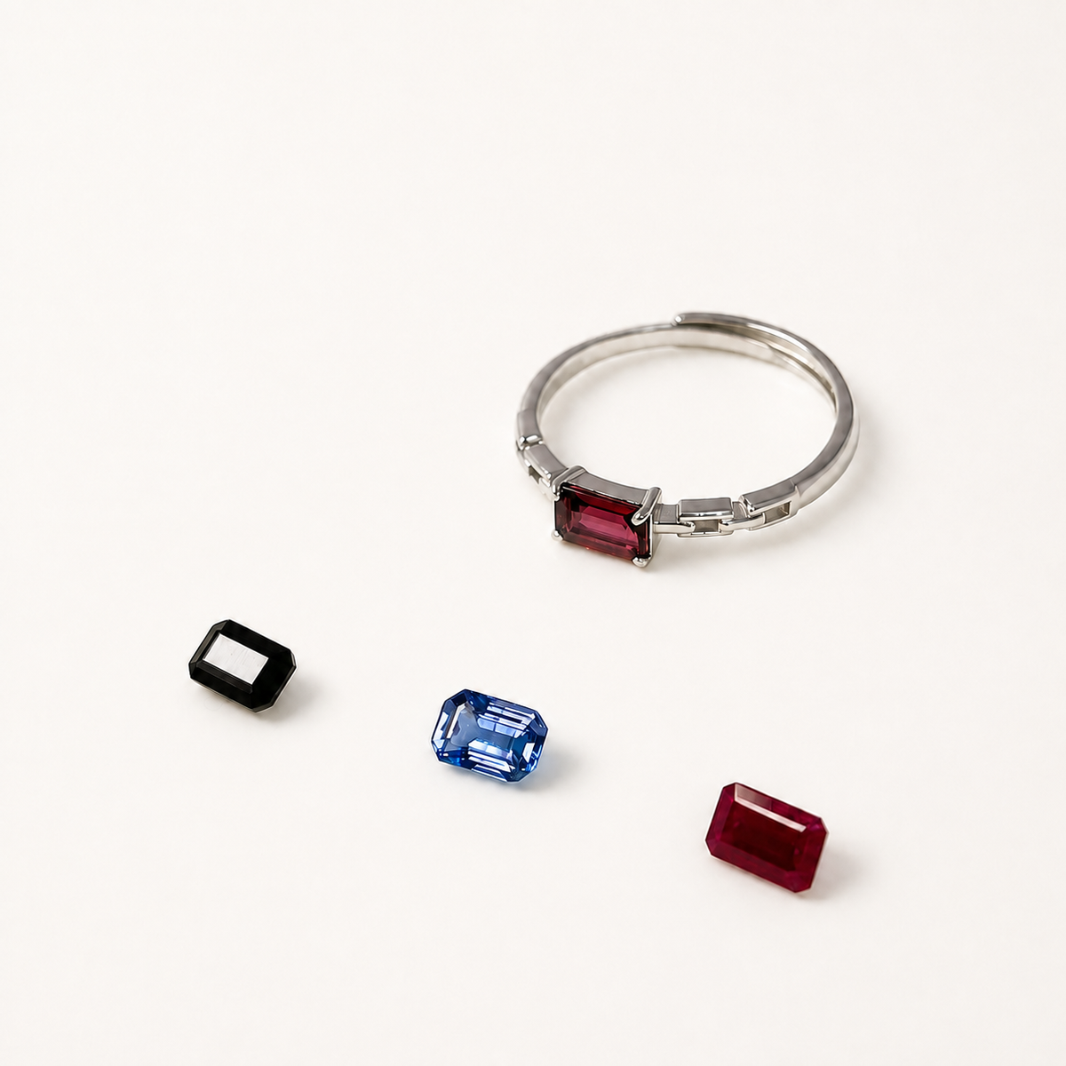 A silver-toned ring with a rectangular red gemstone, shown alongside three loose rectangular gemstones in black, blue, and red on a clean white background.