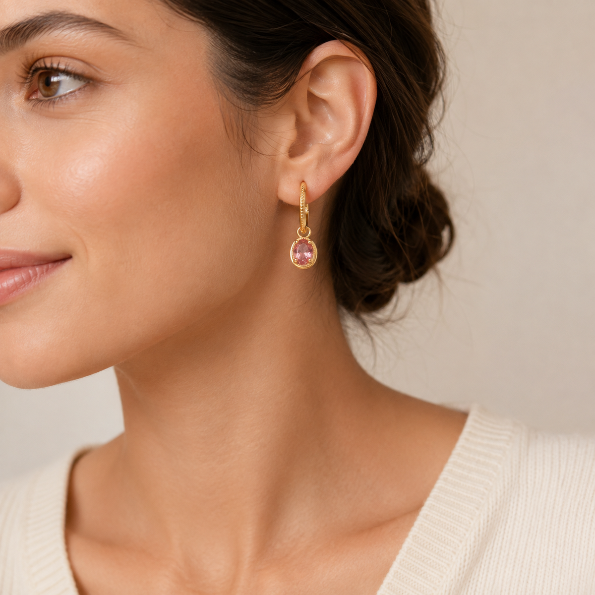 A close-up of a woman wearing a delicate gold huggie hoop earring with a small oval pink gemstone dangling from the side.