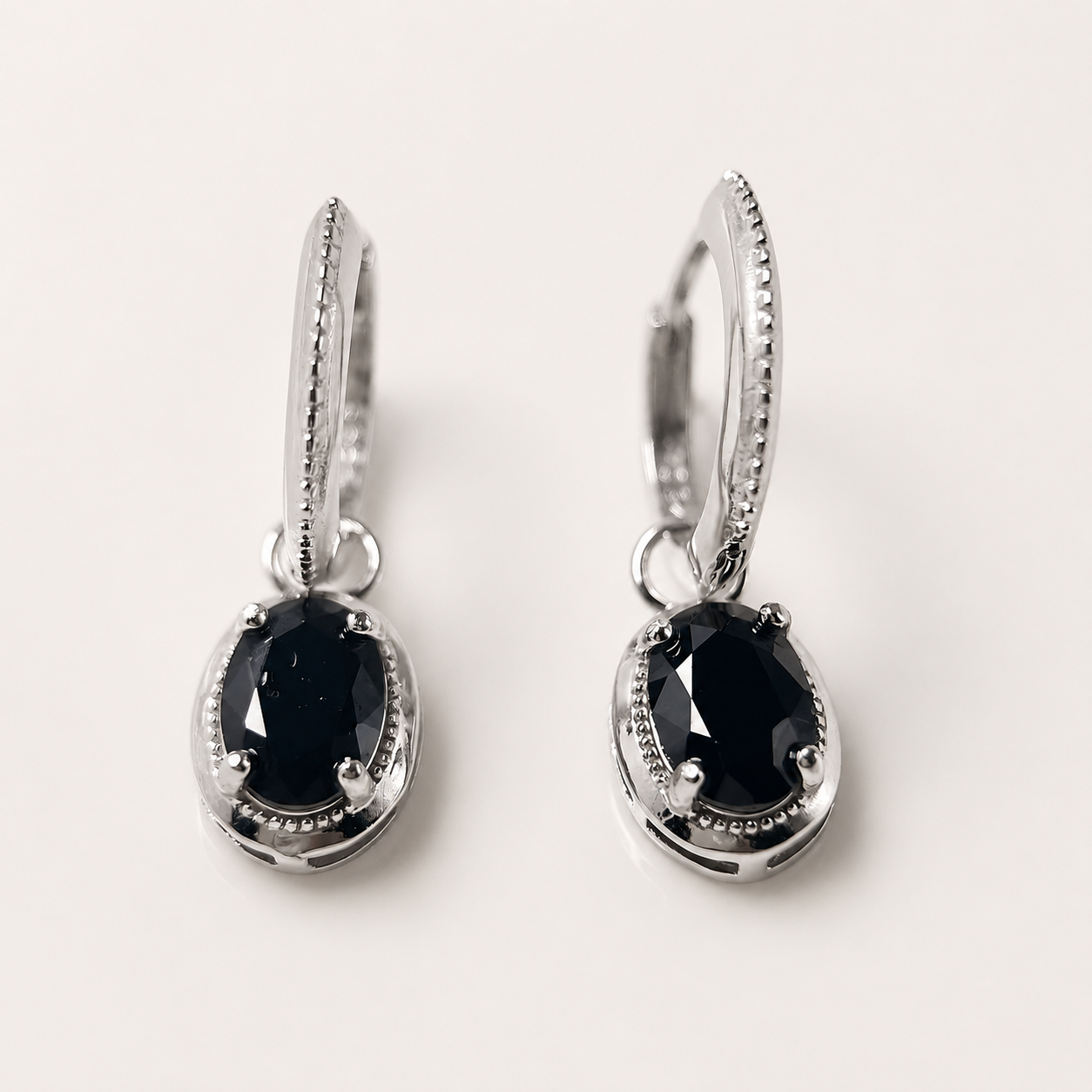 A pair of silver huggie hoop earrings with oval black gemstones, shown on a clean off-white background.