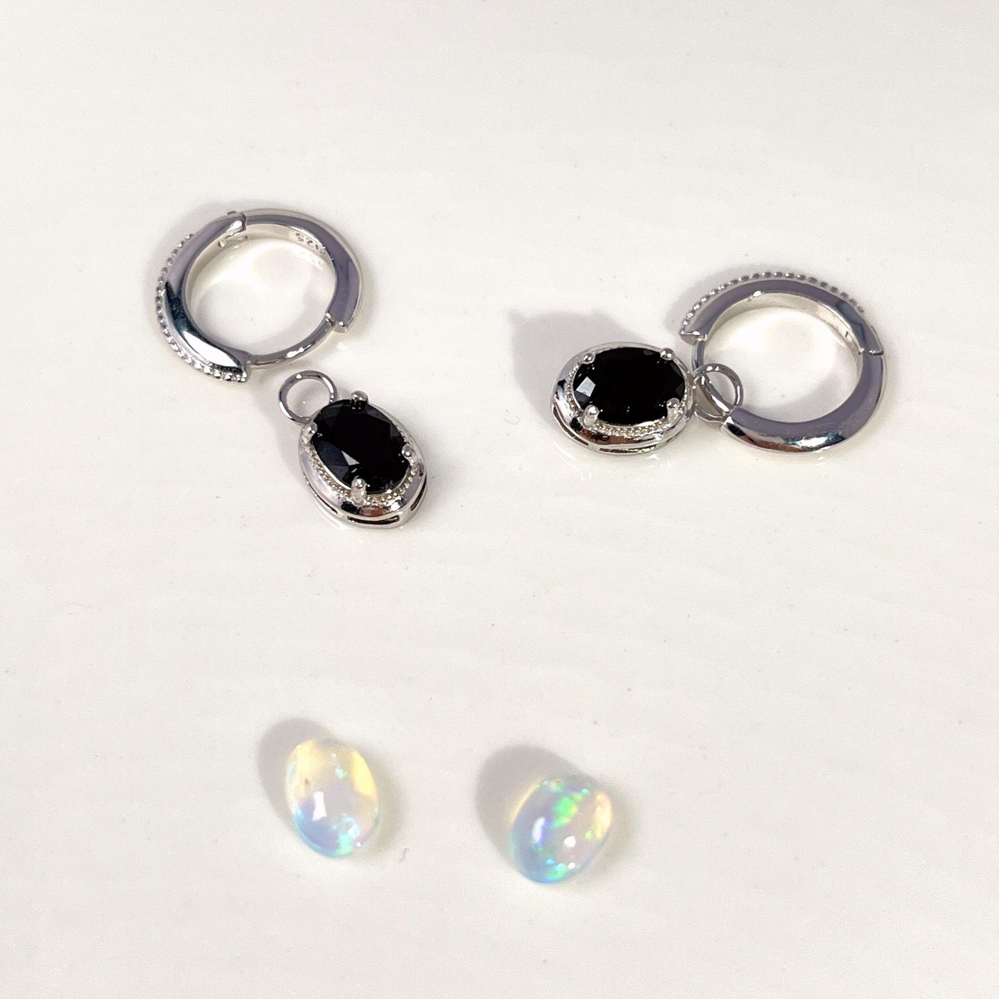 A pair of silver huggie hoop earrings with dangling oval black gemstone charms, displayed alongside two small iridescent white opal-like beads on a plain white background.