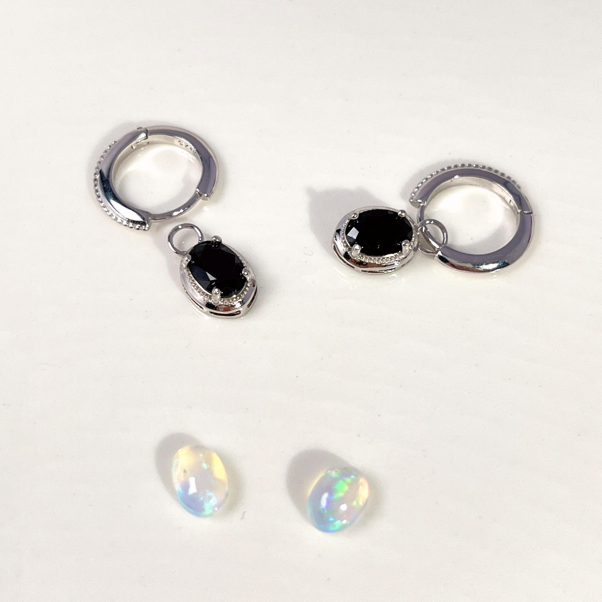A pair of silver huggie hoop earrings with dangling oval black gemstone charms, displayed alongside two small iridescent white opal-like beads on a plain white background.