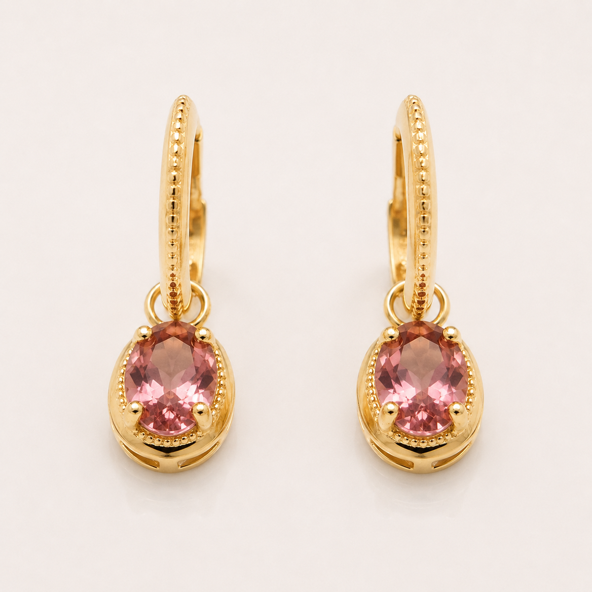 A pair of gold huggie hoop earrings with oval-shaped pink gemstones drop, displayed on a clean white background.
