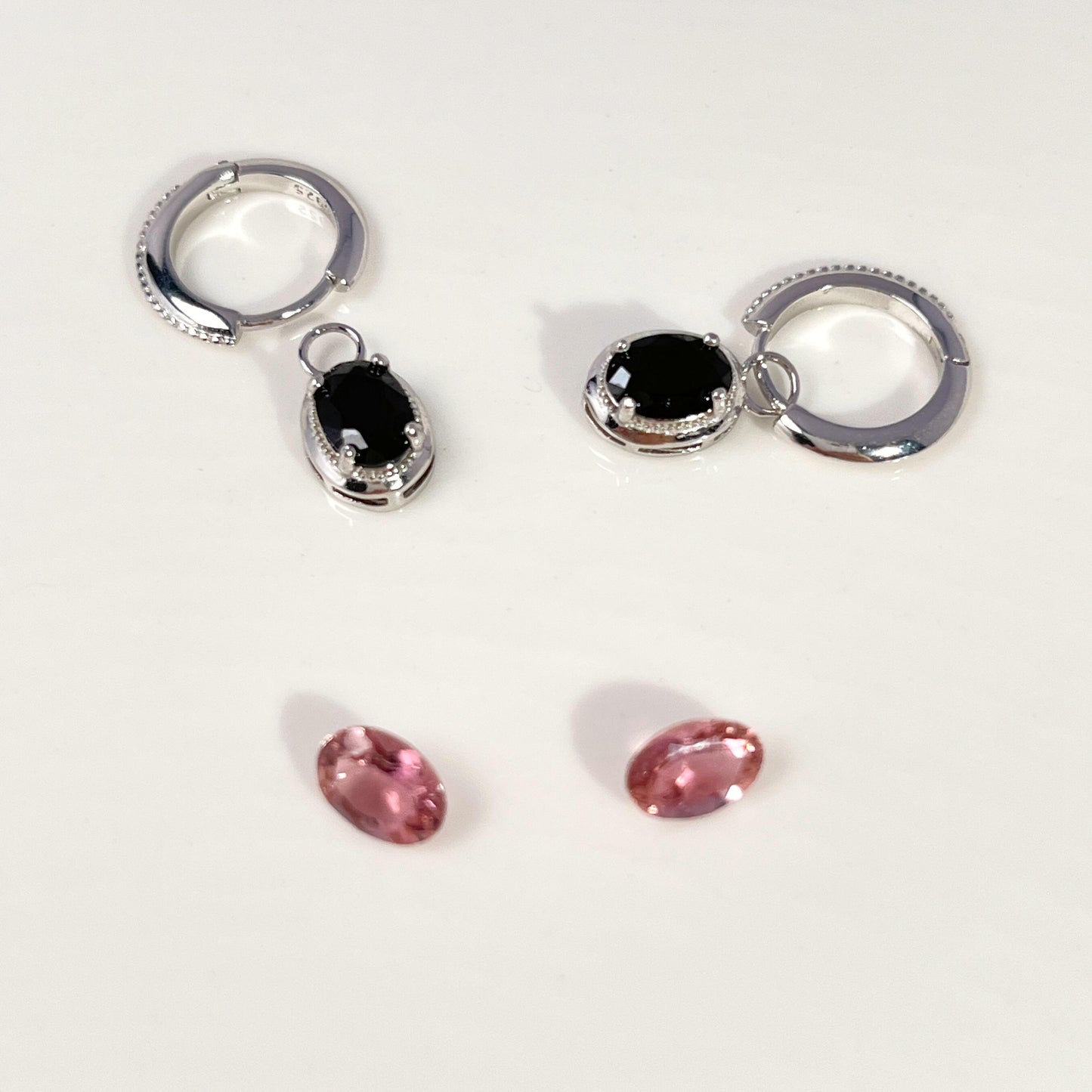 Two silver huggie hoop earrings with oval black gemstones, shown alongside two small oval pink gemstones on a plain white background.