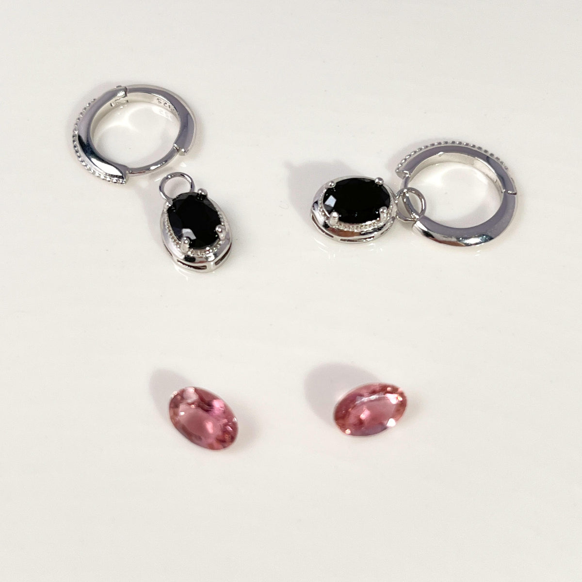 Two silver huggie hoop earrings with oval black gemstones, shown alongside two small oval pink gemstones on a plain white background.