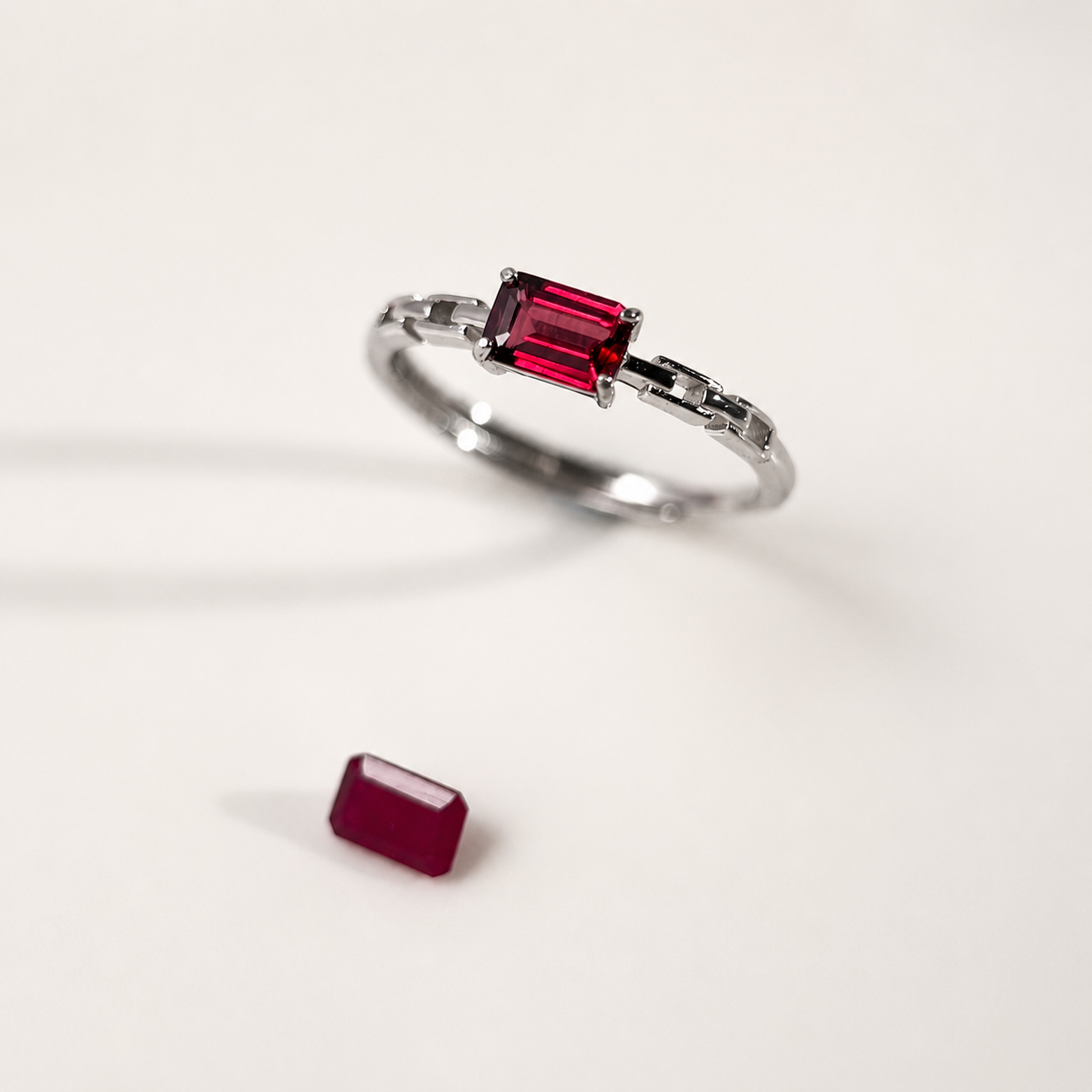 A silver ring featuring a rectangular emerald-cut purple-red gemstone, shown on a clean white background with a small, loose ruby placed below it.