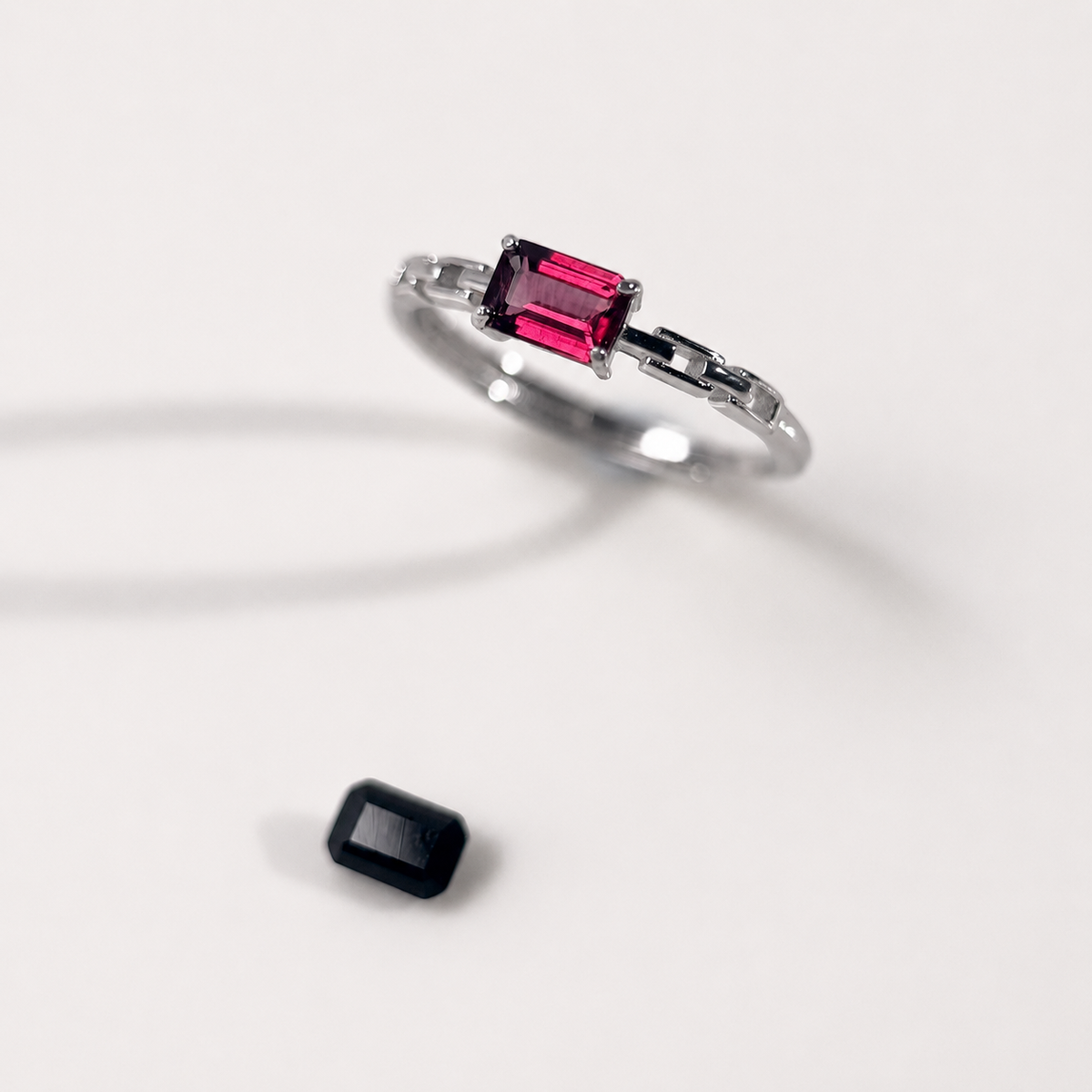 A silver ring featuring a rectangular emerald-cut purple-red gemstone, shown on a clean white background with a small matching loose gemstone placed below it.