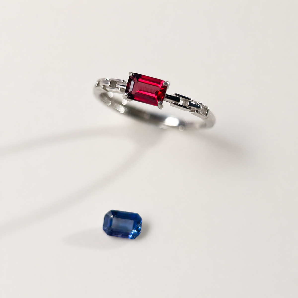 A silver ring featuring an emerald-cut purple-red gemstone, shown on a light gray background with a separate blue emerald-cut gemstone placed below it.