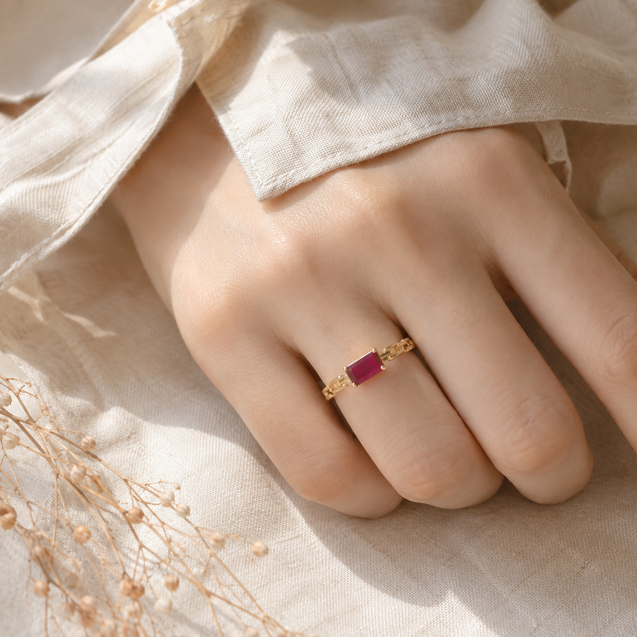 A gold ring with a rectangular ruby worn on a person's finger, set against a soft beige fabric background with dried flowers.