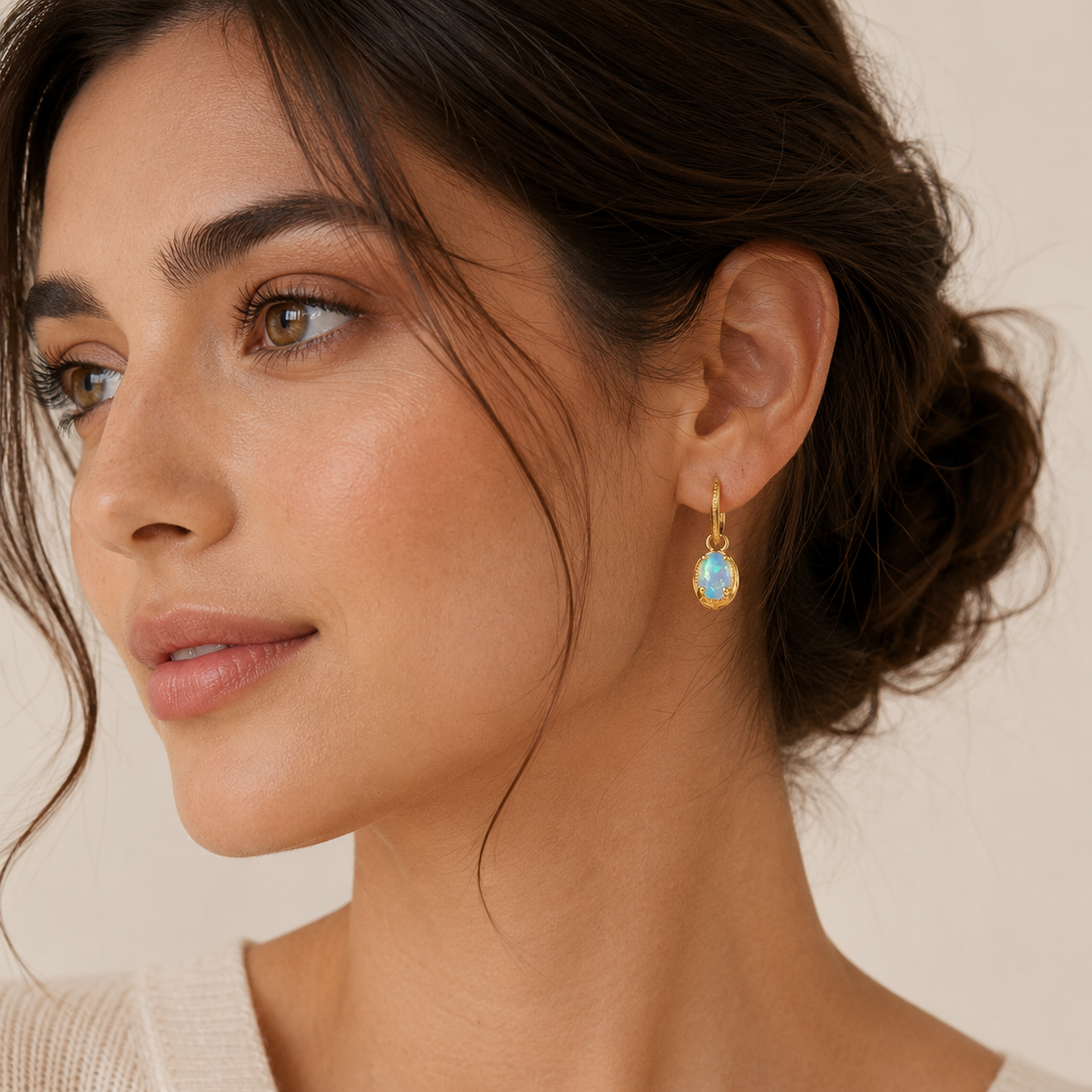 A close-up of a woman wearing a delicate gold dangle earring with a oval-shaped light blue opal stone drop, set against a warm beige background.