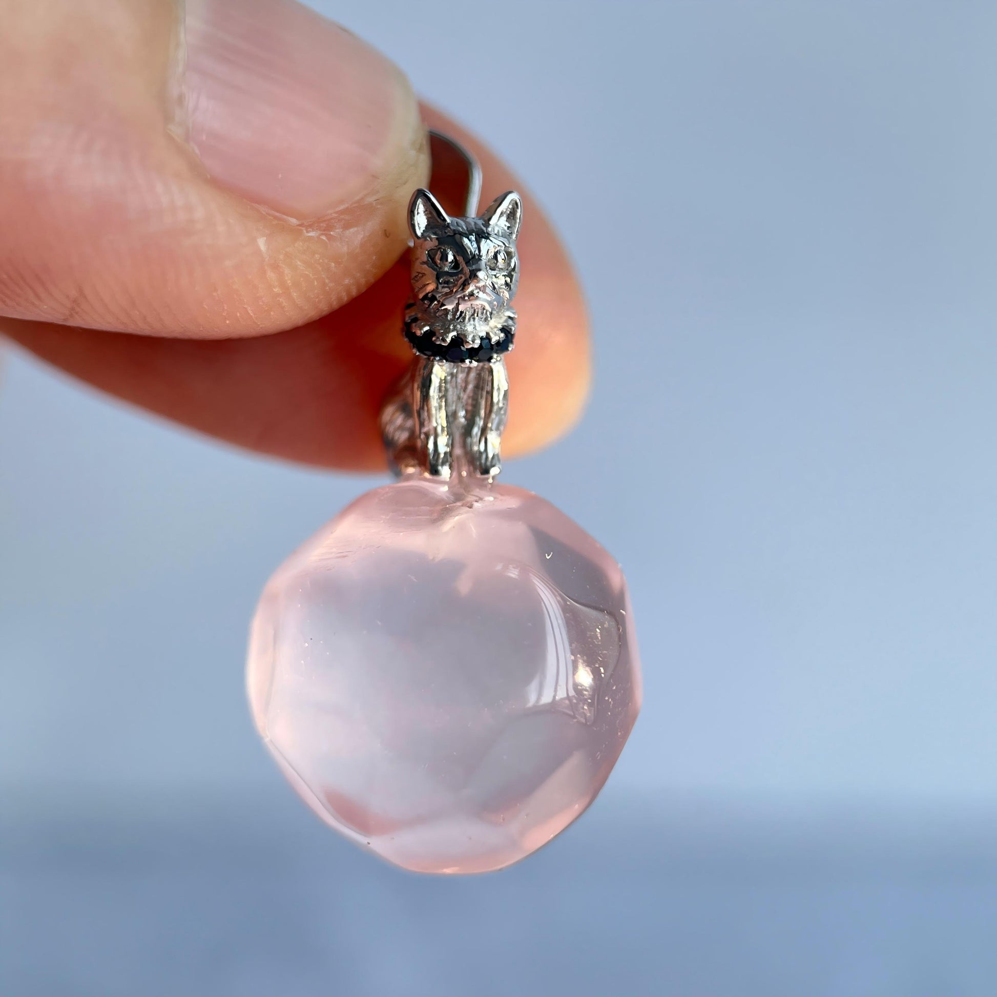 Pink crystal pendant with silver cat-shaped top held by a hand against a light blue background