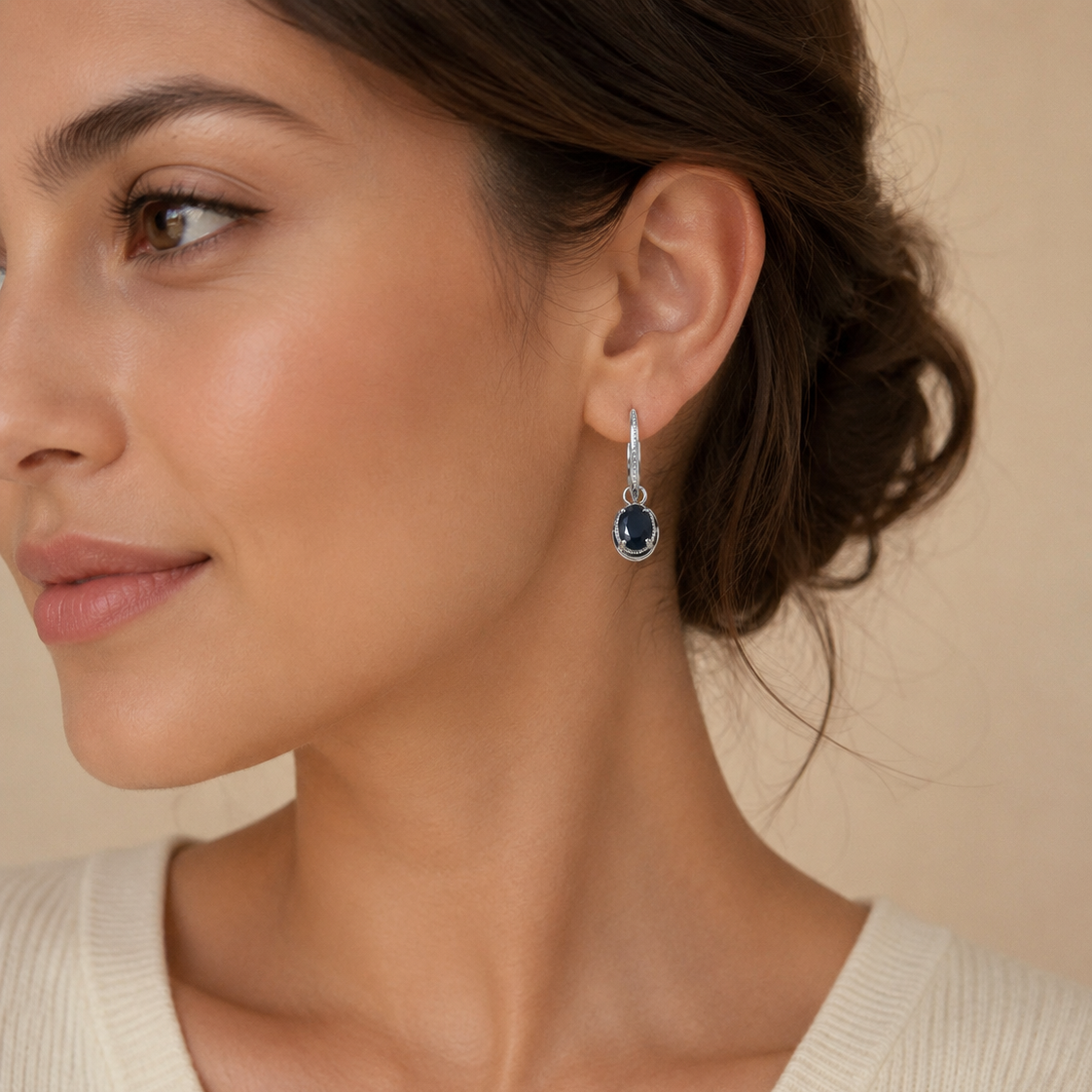 A close-up of a woman wearing a silver drop earring with a dark oval gemstone, set against a soft beige background.