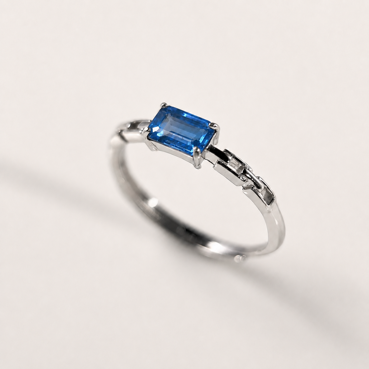 A silver-toned ring featuring a rectangular blue gemstone in a prong setting, photographed on a clean off-white background.