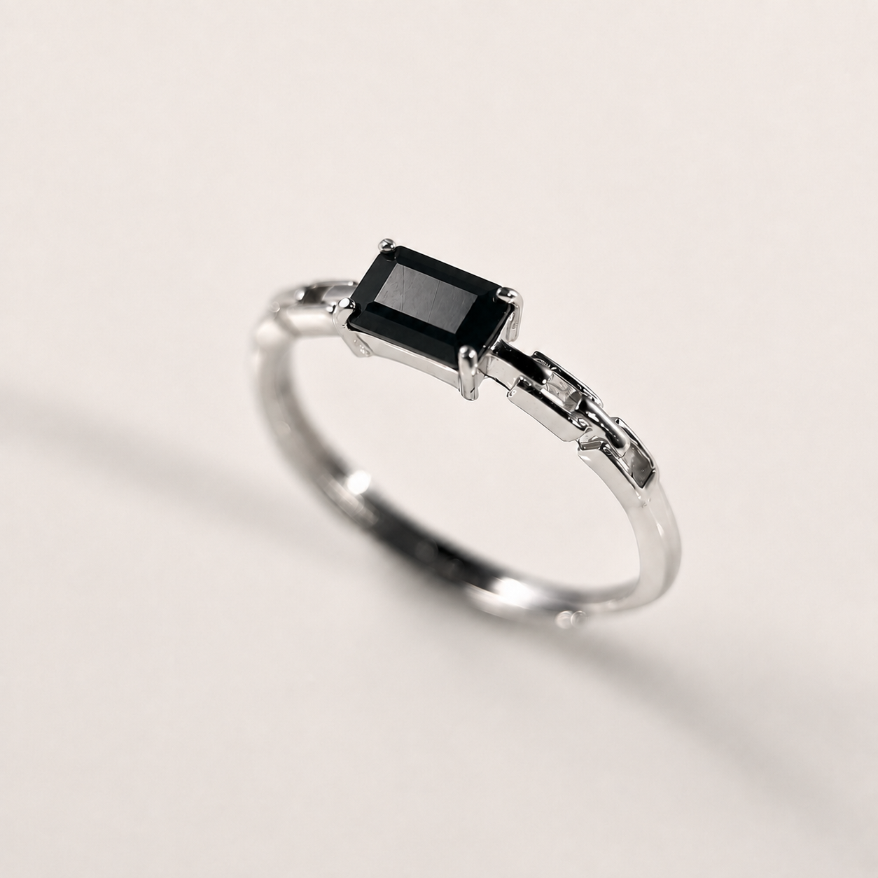 A silver ring featuring a rectangular black gemstone in a prong setting, shown on a clean, light beige background.