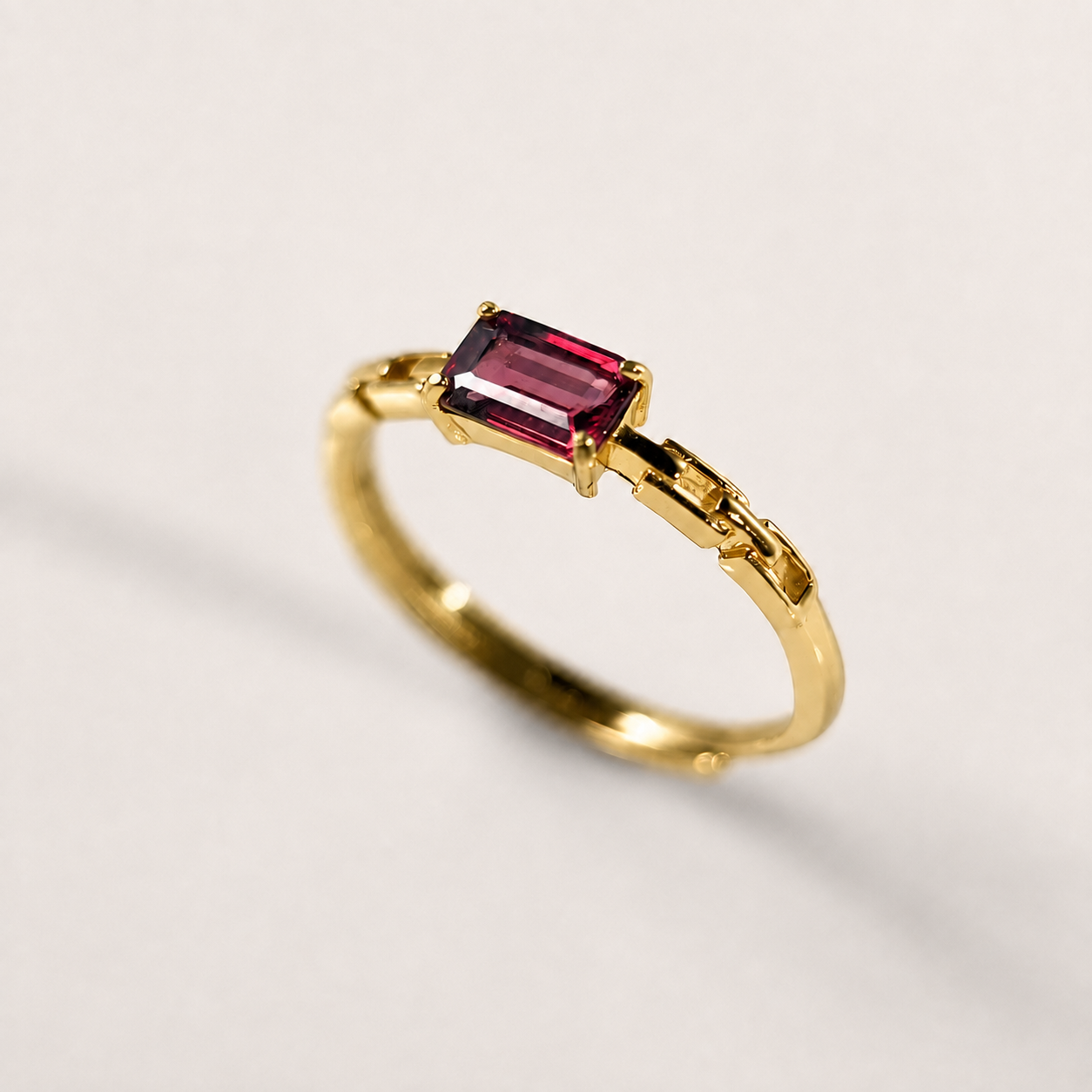 A gold ring featuring a rectangular emerald-cut pinkish-red gemstone, photographed on a clean, light off-white background.