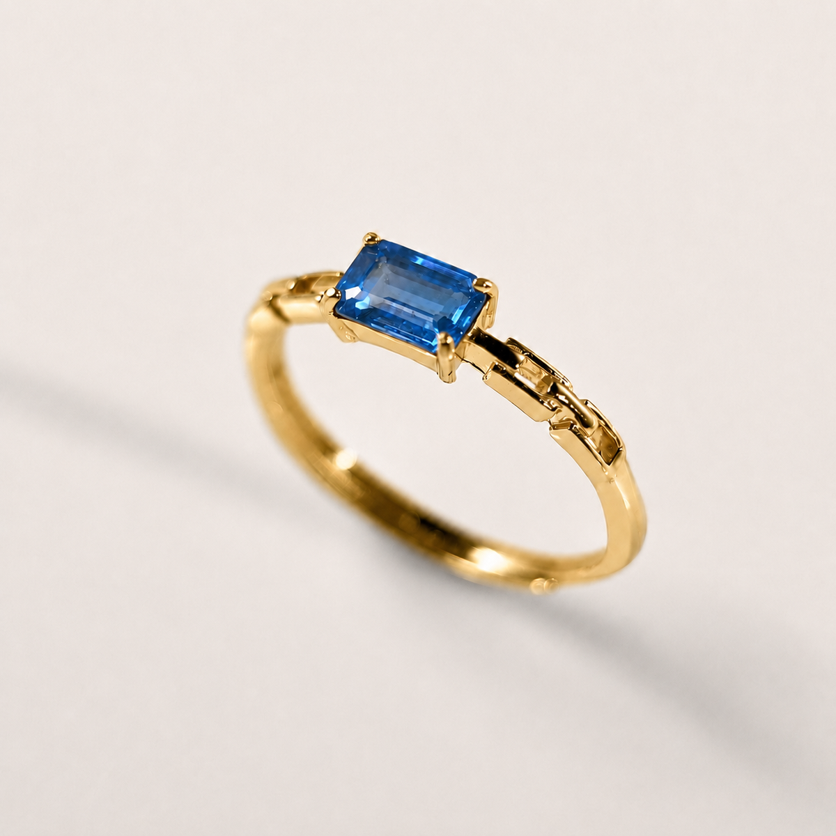 A gold ring featuring a rectangular blue gemstone in a prong setting, shown on a clean, light off-white background.
