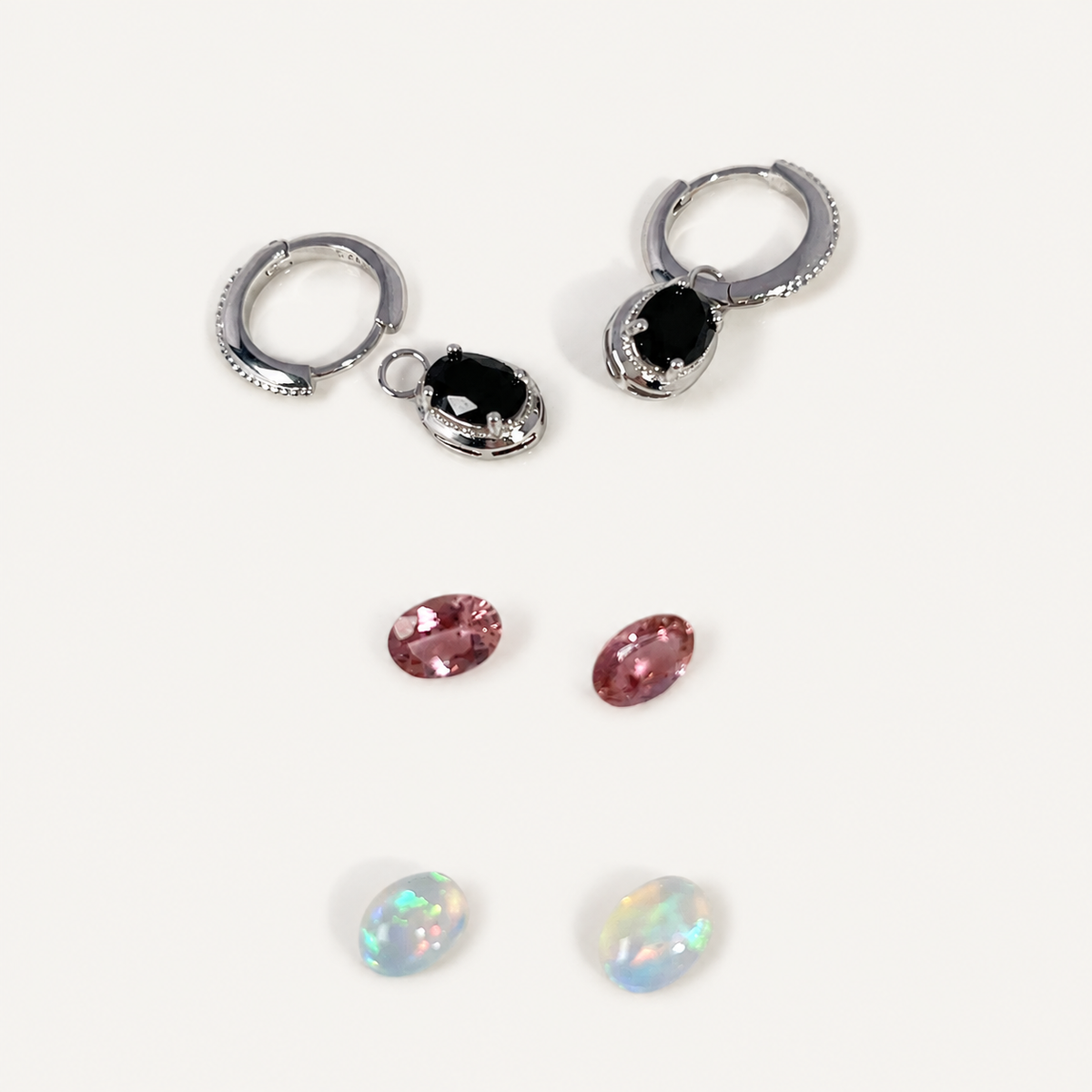 Silver hoop earrings with oval black faceted gemstones, shown alongside four loose oval gemstones in pink, pink, and iridescent white-blue opal-like colors on a plain white background.