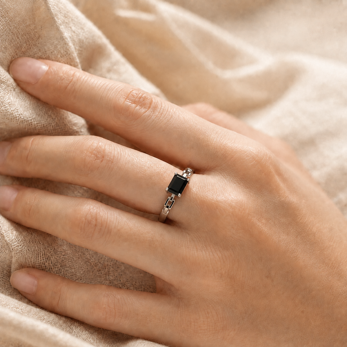 A close-up of a hand wearing a silver ring with a rectangular black gemstone, resting on a soft beige fabric background.