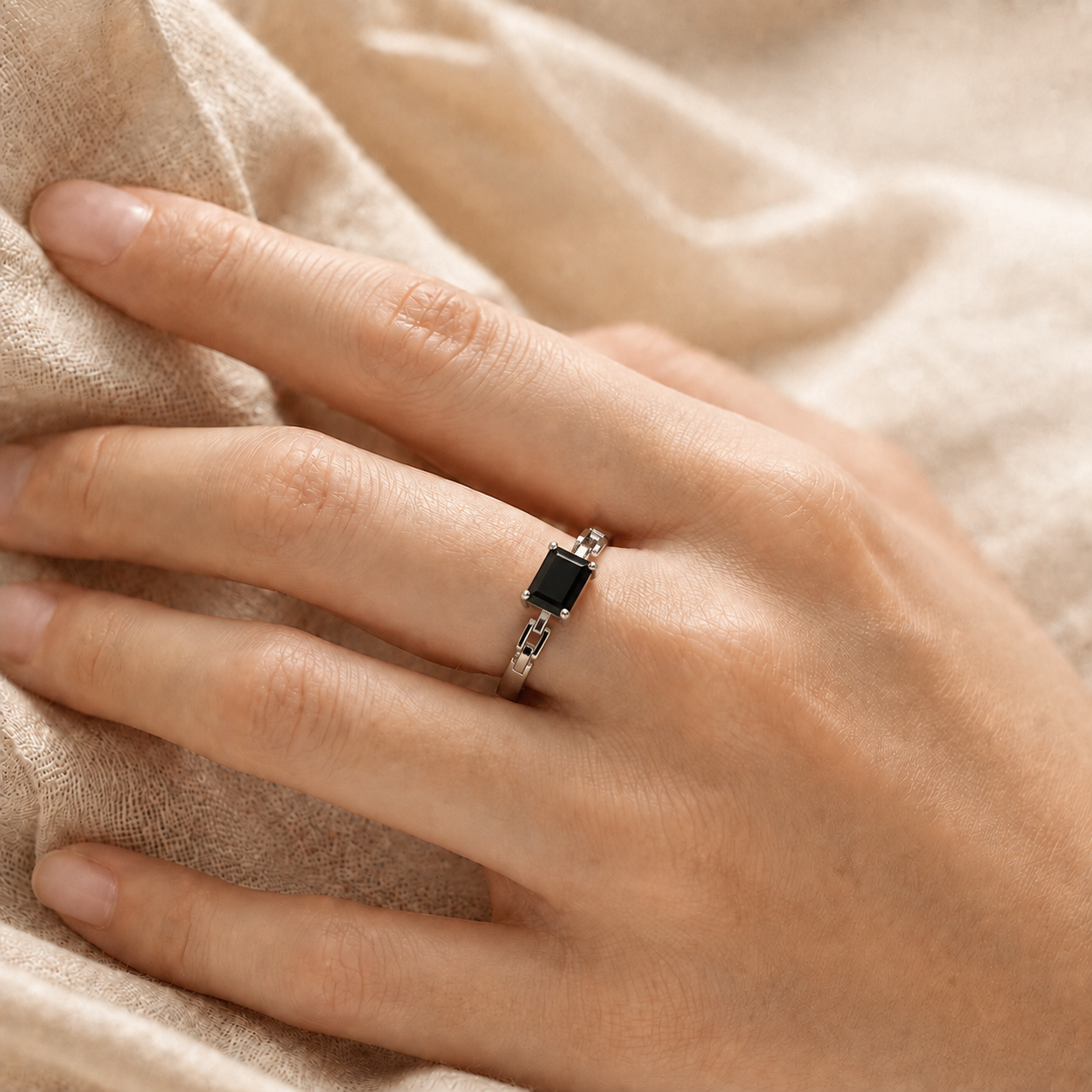 A close-up of a hand wearing a silver ring with a rectangular black gemstone, resting on a soft beige fabric background.