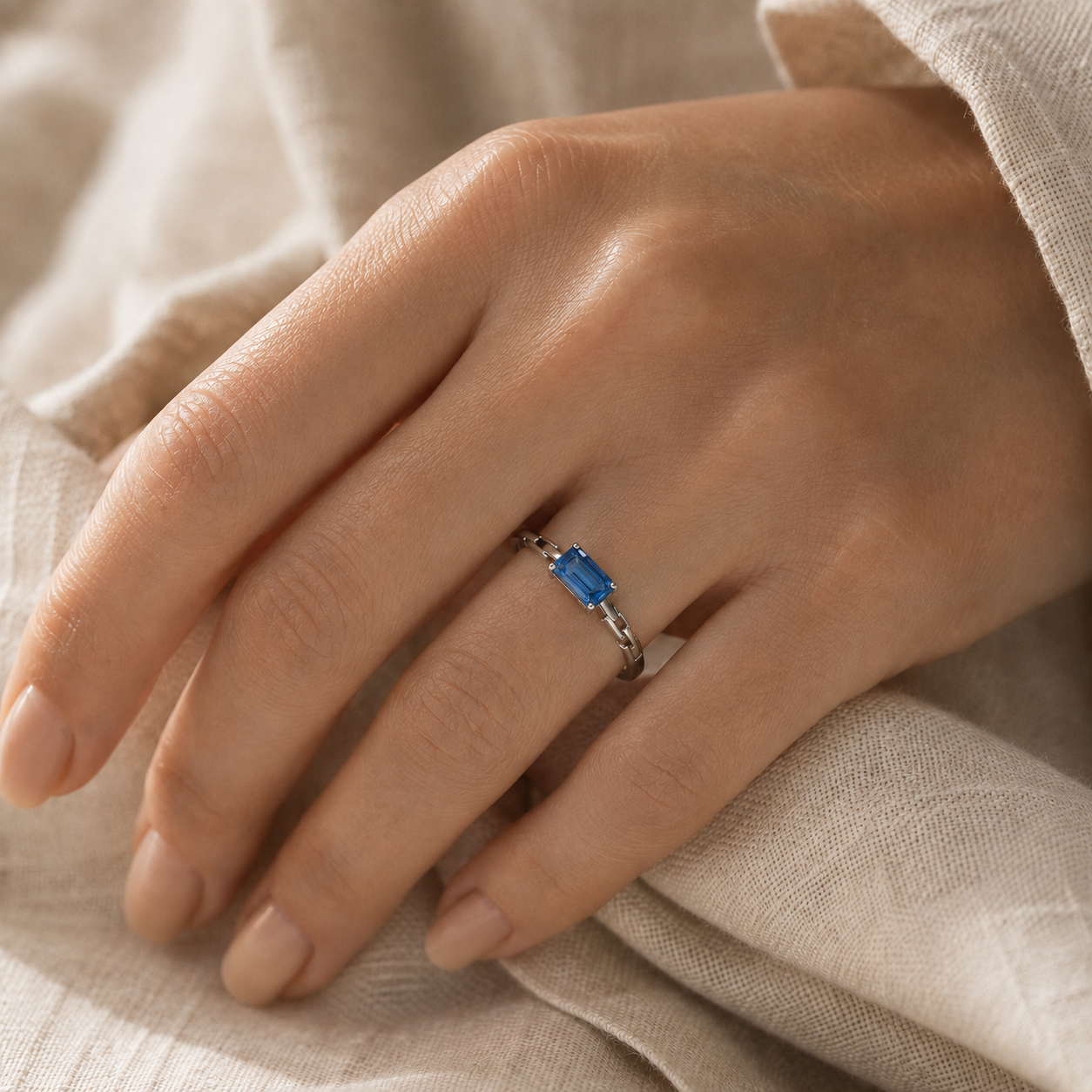 A close-up of a hand wearing a silver ring with a rectangular blue gemstone, resting on a soft beige fabric background.