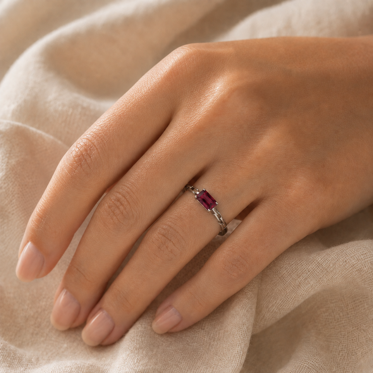 A close-up of a hand wearing a silver ring with a rectangular red gemstone, resting on soft beige fabric.