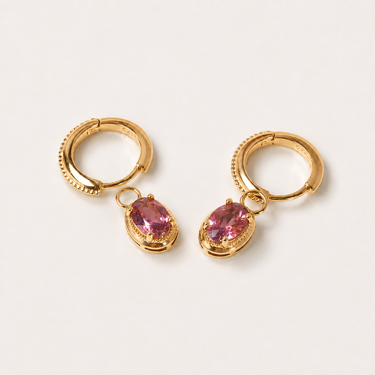 A pair of gold huggie hoop earrings with dangling oval pink gemstones, displayed on a clean off-white background.