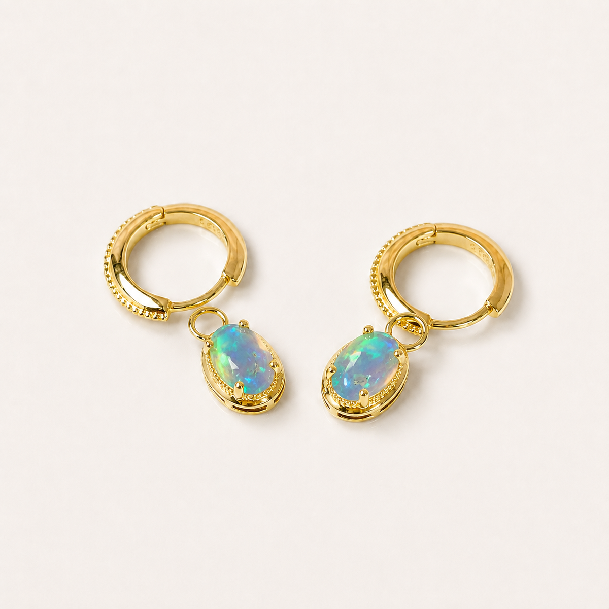 A pair of gold huggie hoop earrings with dangling oval-shaped opal gemstones, displayed on a clean off-white background.
