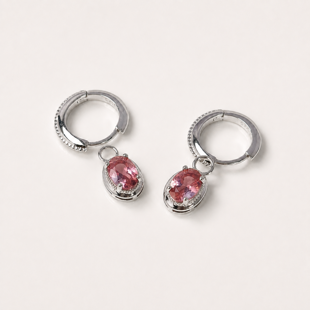A pair of silver huggie hoop earrings with dangling oval pink gemstone charms, displayed on a clean off-white background.
