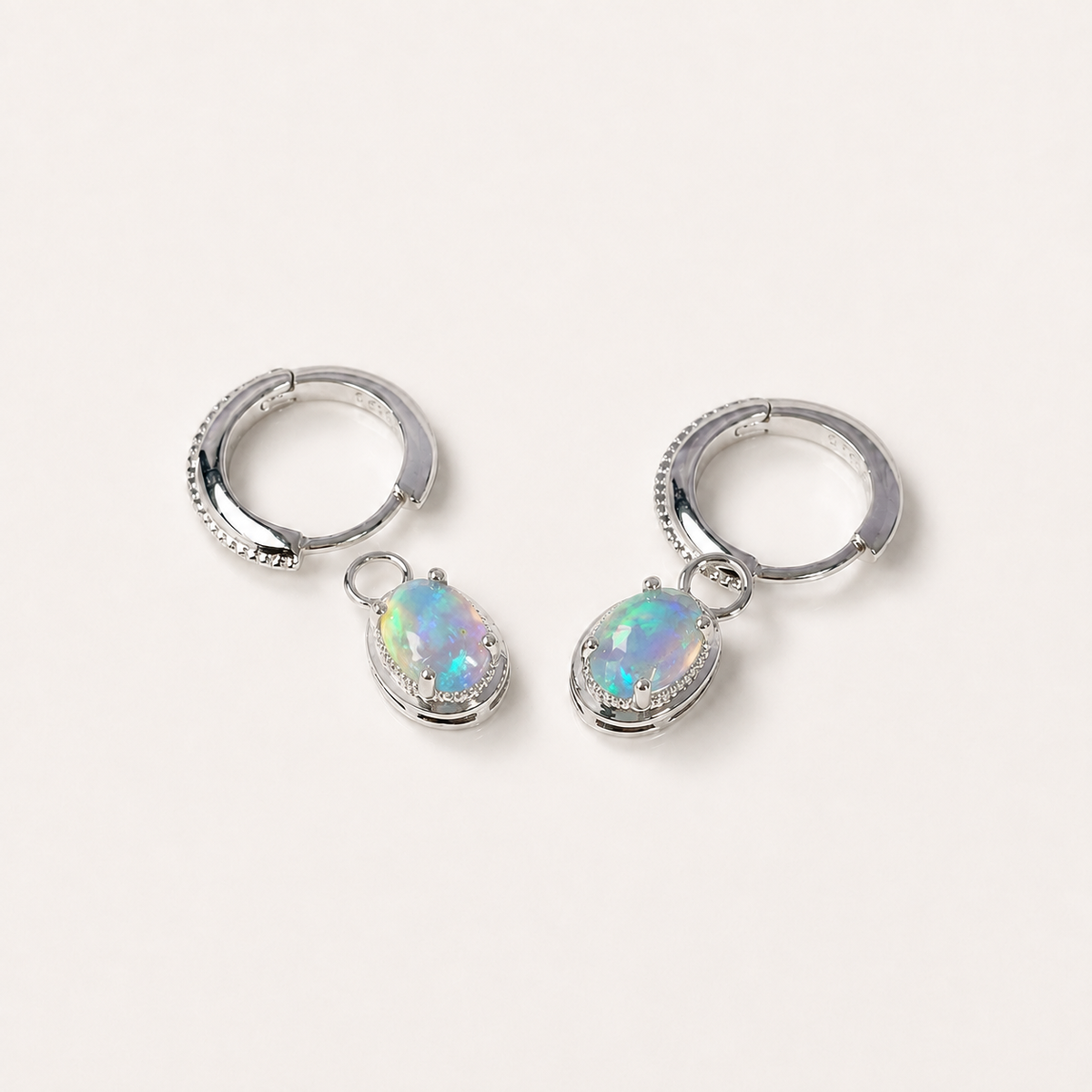 A pair of silver huggie hoop earrings with dangling oval-shaped opal gemstones, displayed on a clean off-white background.