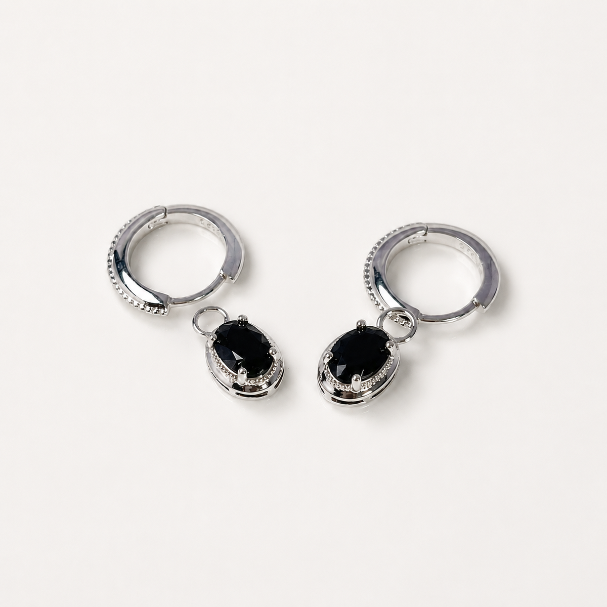 A pair of silver hoop earrings with dangling oval black gemstone charms, photographed on a clean, light off-white background.
