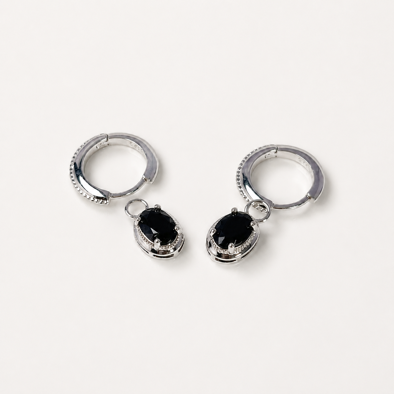 A pair of silver hoop earrings with dangling oval black gemstone charms, photographed on a clean, light off-white background.