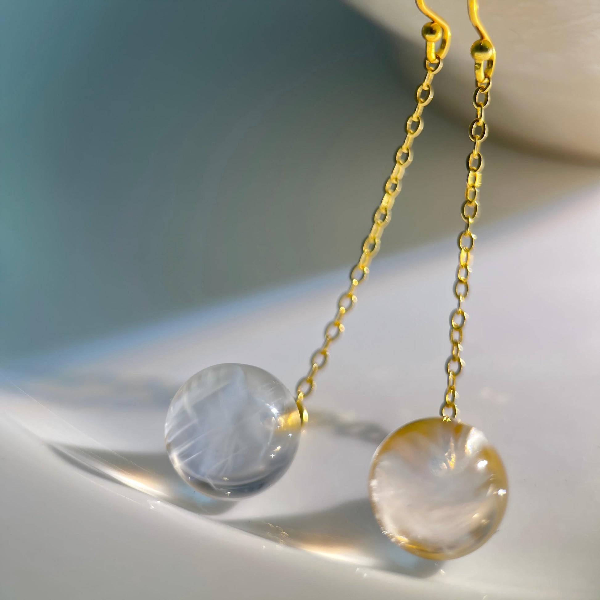 A pair of gold chain drop earrings with round clear gemstone pendants, resting on a glossy white surface with soft shadows.