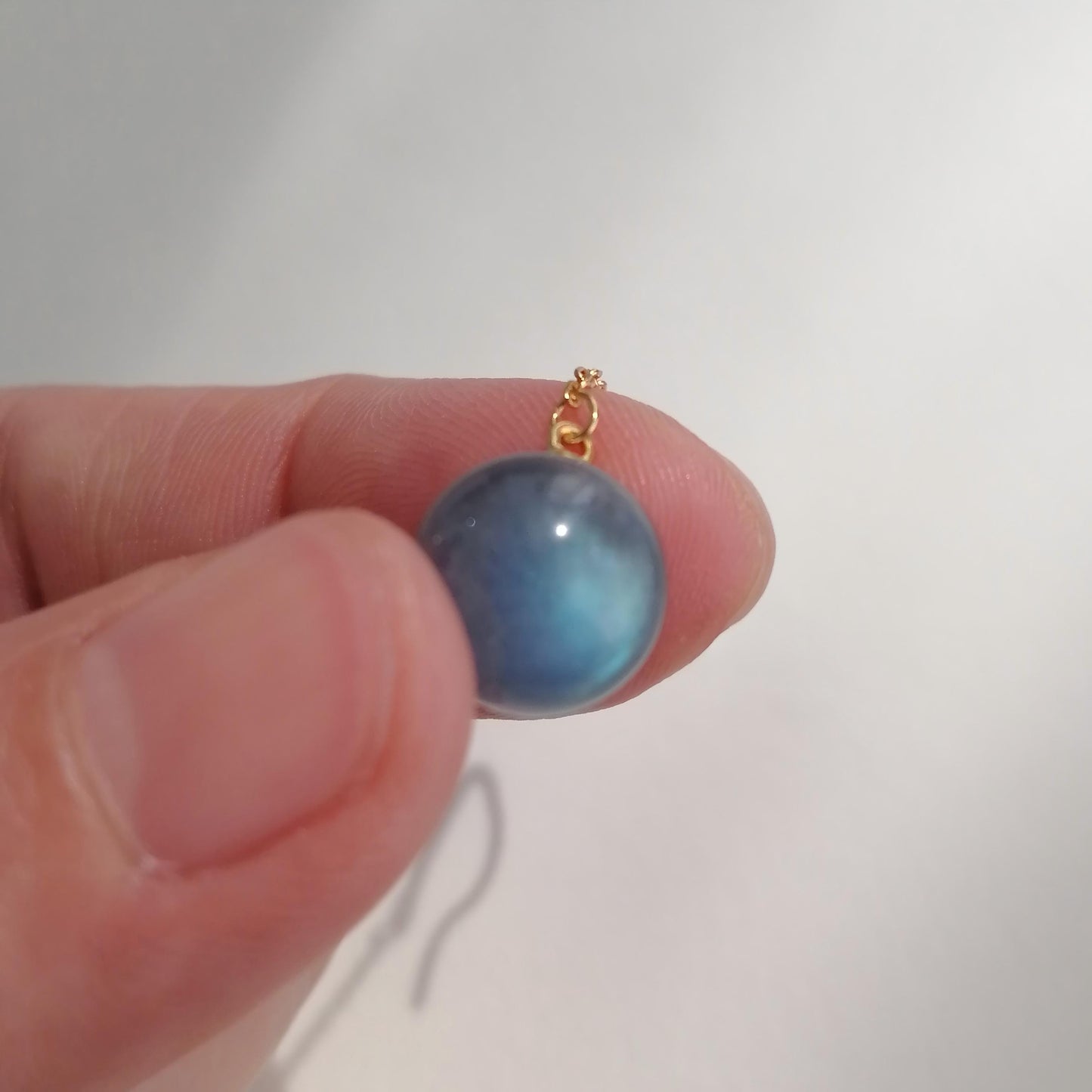 Blue cat's eye gemstone on a gold chain held between fingers against a white background