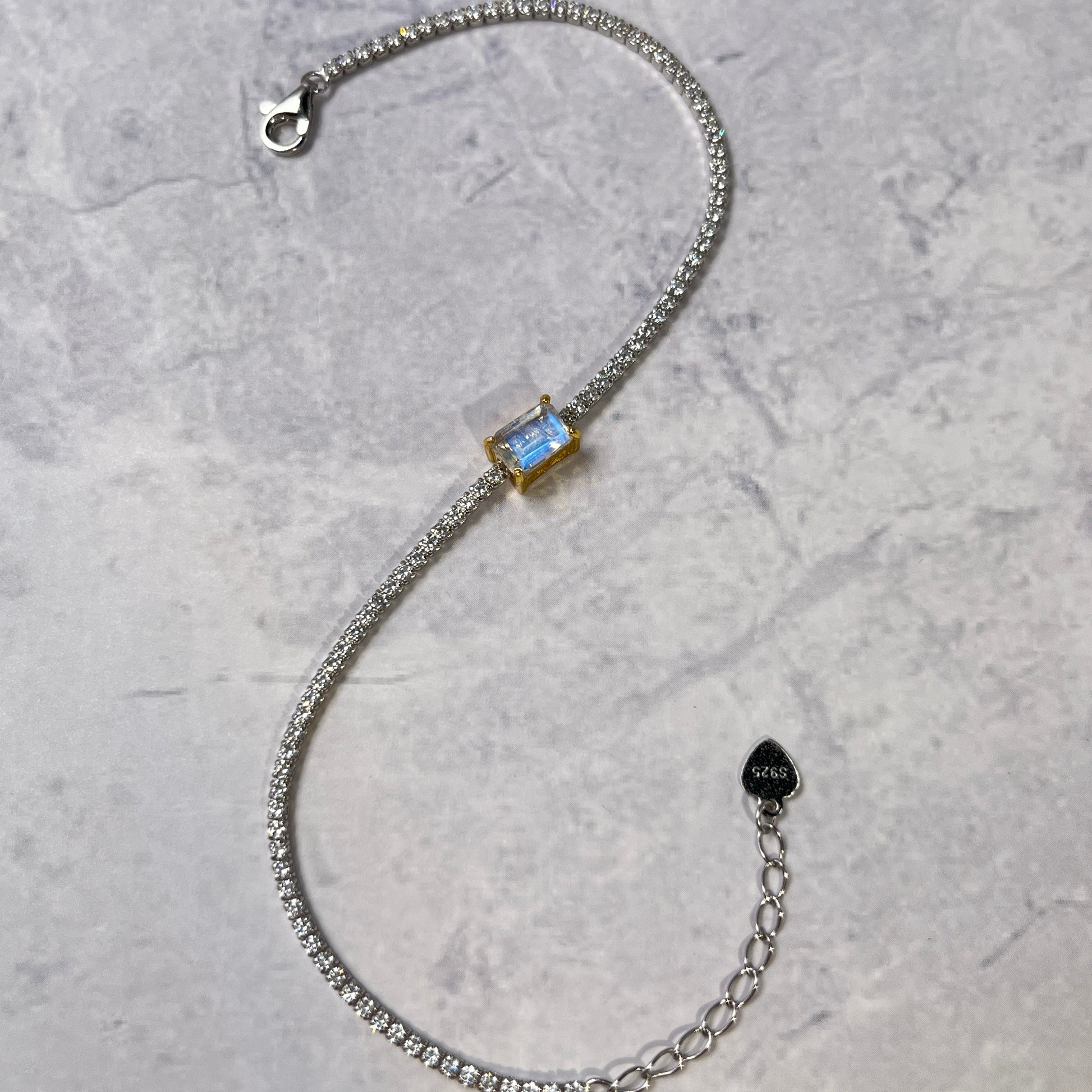A silver chain bracelet with a rectangular light blue gemstone centerpiece, displayed on a textured gray background. The bracelet features a lobster clasp, an adjustable extension chain with a small heart-shaped tag, and a bezel-set blue stone.