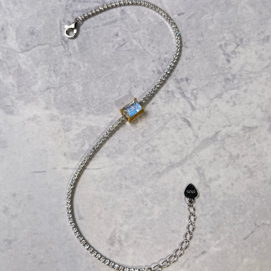 A silver chain bracelet with a rectangular light blue gemstone centerpiece, displayed on a textured gray background. The bracelet features a lobster clasp, an adjustable extension chain with a small heart-shaped tag, and a bezel-set blue stone.