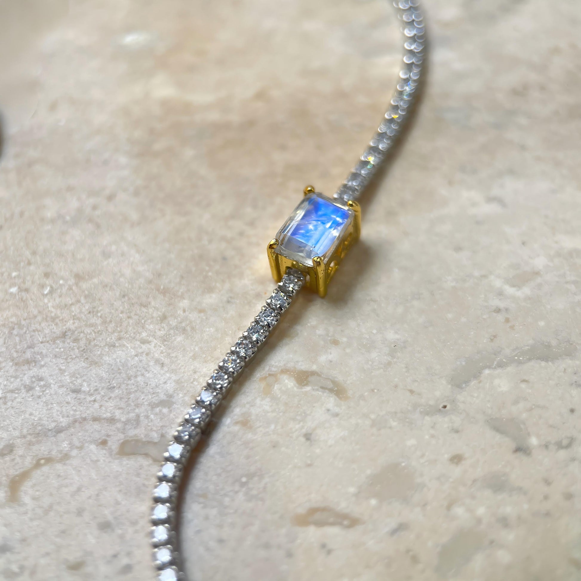 A close-up of a delicate silver pavé chain necklace with a central rectangular emerald-cut iridescent blue stone in a gold setting, resting on a light beige stone surface.