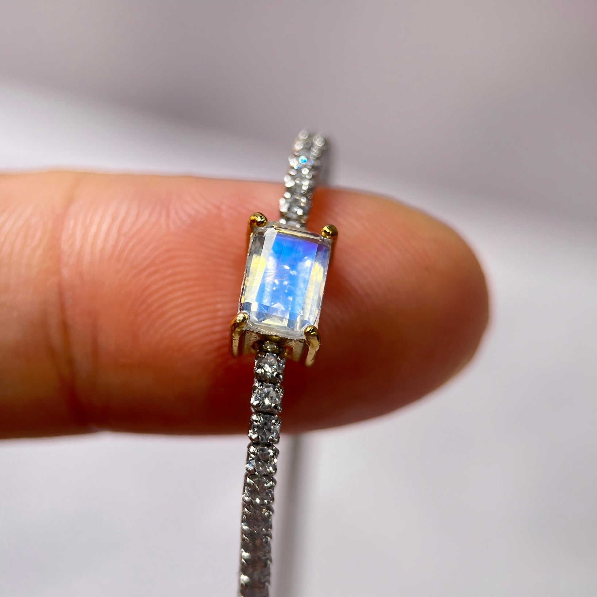 A close-up of a ring held between fingers, featuring a rectangular iridescent blue-white center stone in a gold setting with a pavé band of clear stones on a white background.
