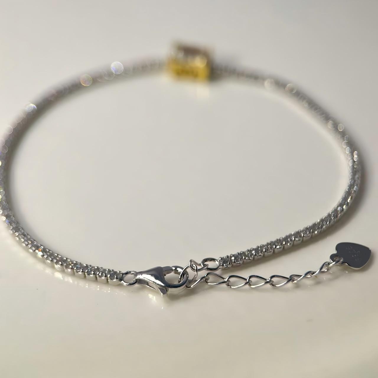 A silver chain bracelet with a lobster clasp, extension chain, and a small heart-shaped tag, displayed on a light beige surface.