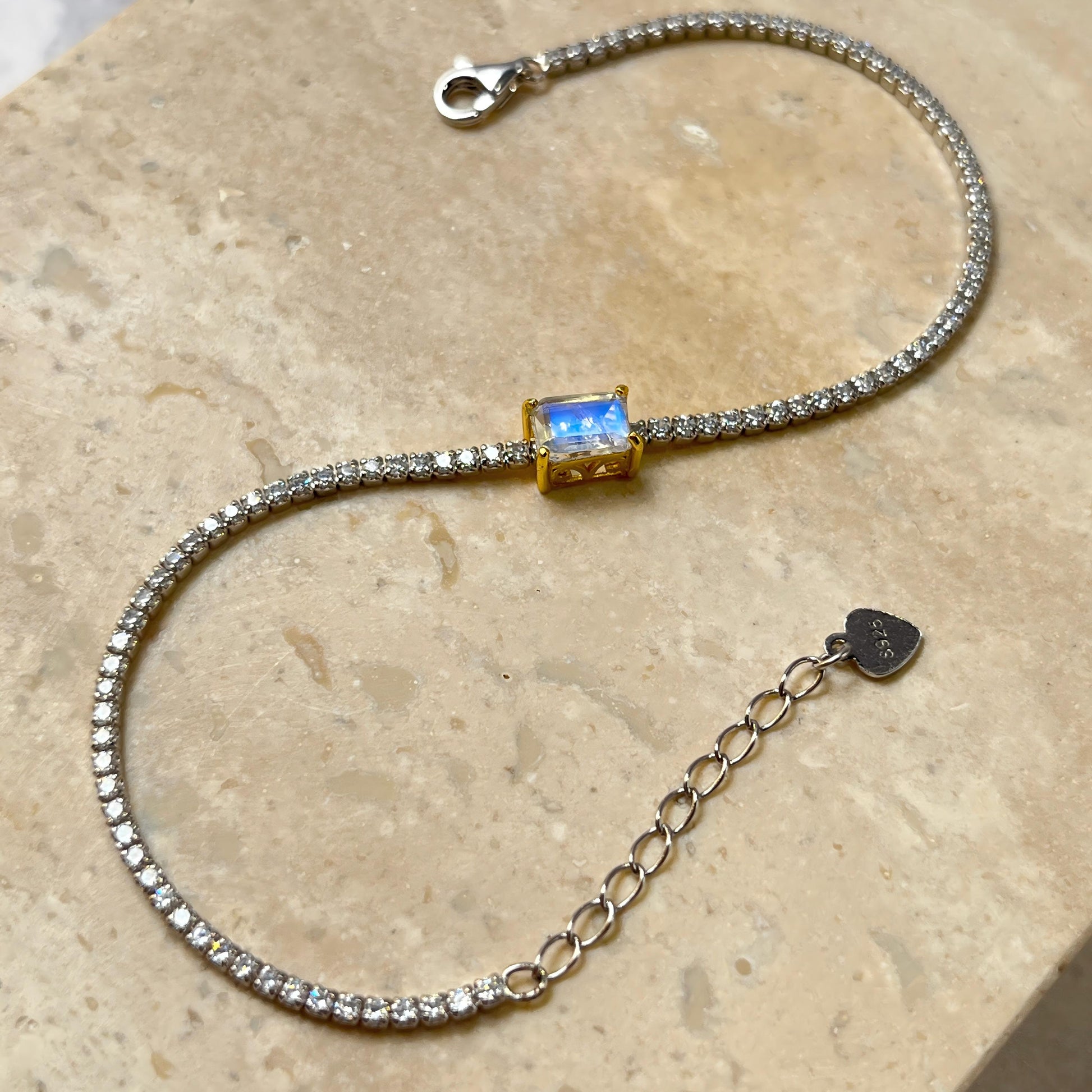 A silver tennis bracelet paved with small clear stones, featuring a central gold square bezel with a blue iridescent gemstone. The bracelet is displayed on a beige stone surface with an adjustable silver chain extender and a small heart-shaped tag.