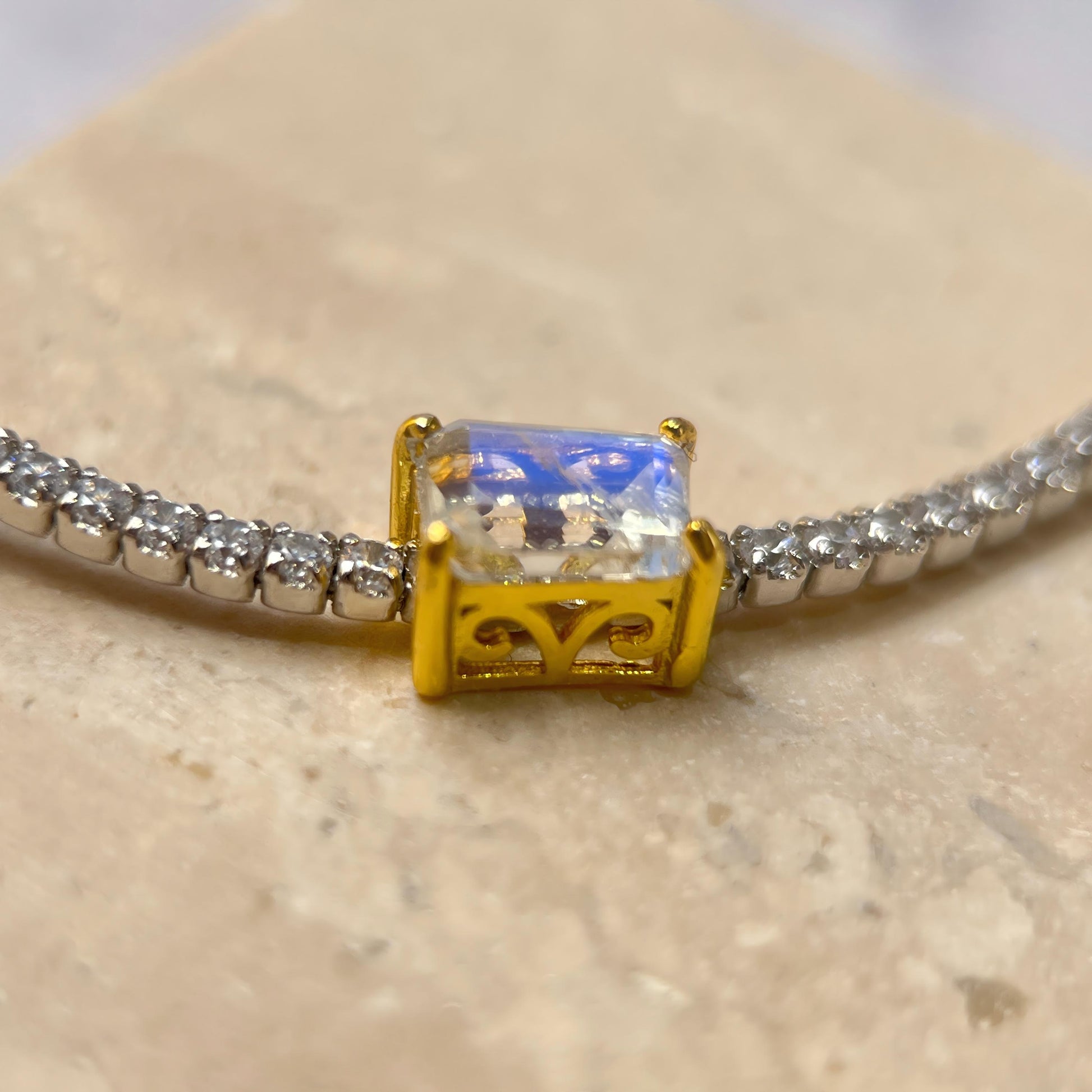 A close-up of a bracelet featuring a rectangular clear gemstone in a gold bezel setting, with a silver chain extending on both sides, resting on a light beige stone surface.
