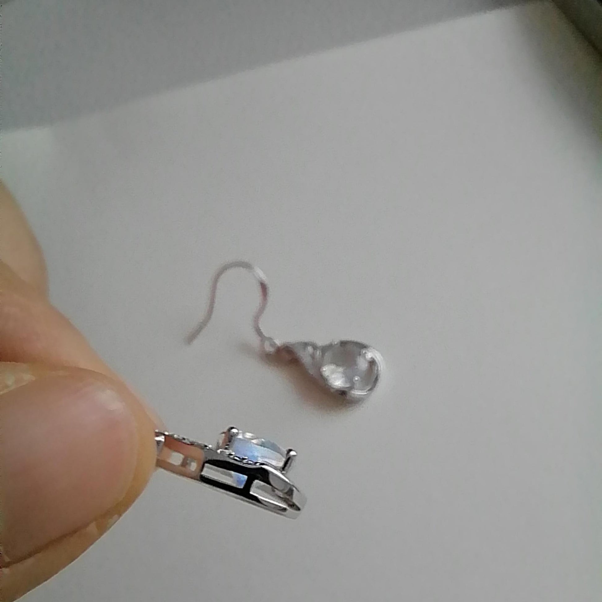 Close-up of a silver earring with a clear gemstone held by a hand against a plain background.