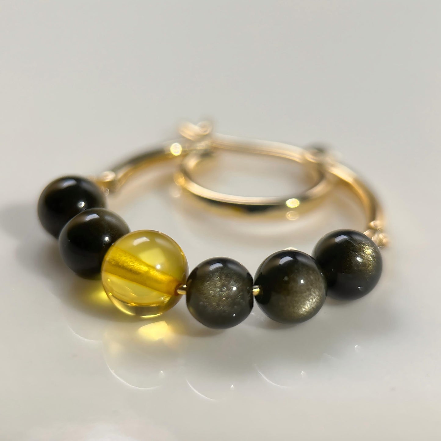 A gold-plated beaded hoop earing featuring six round beads: five dark black beads and one translucent yellow-green bead, displayed on a clean white surface.