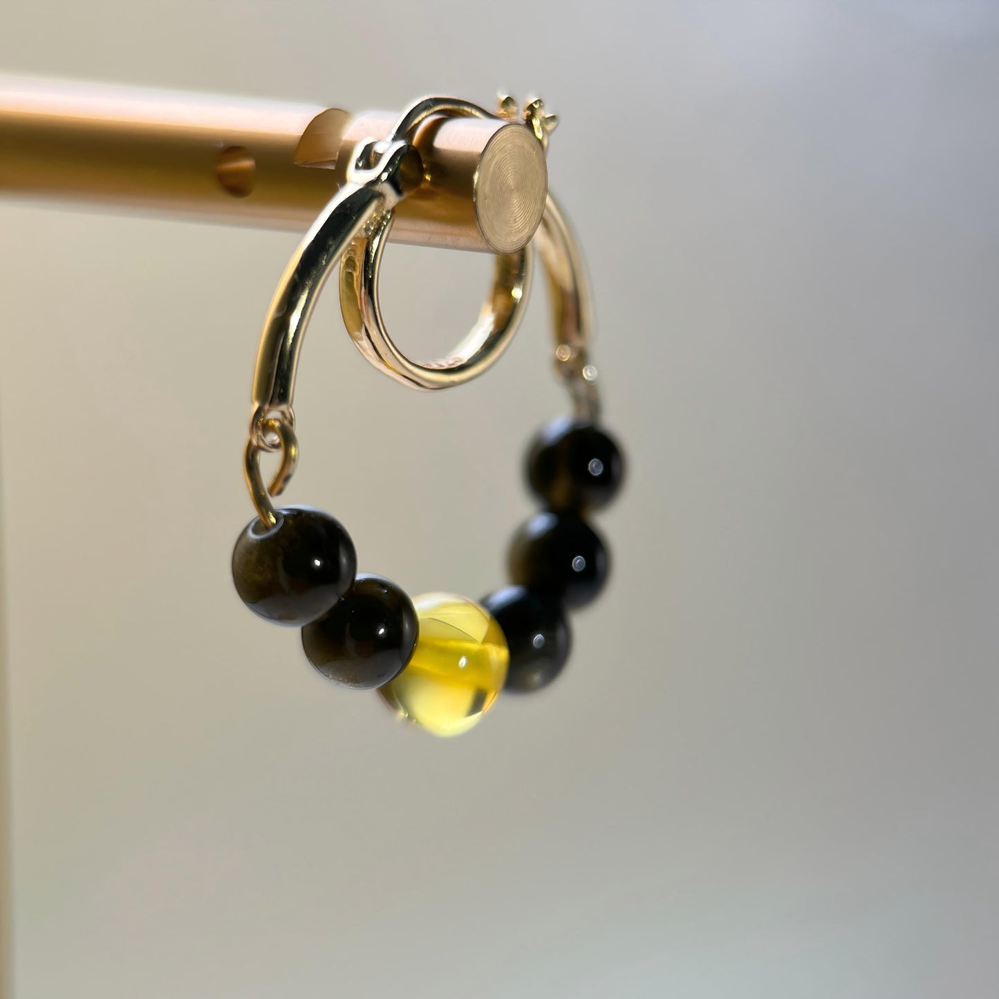 A close-up of a gold hoop earring with a dangling beaded charm featuring black and translucent yellow beads, hanging from a gold display rod against a soft gray background.