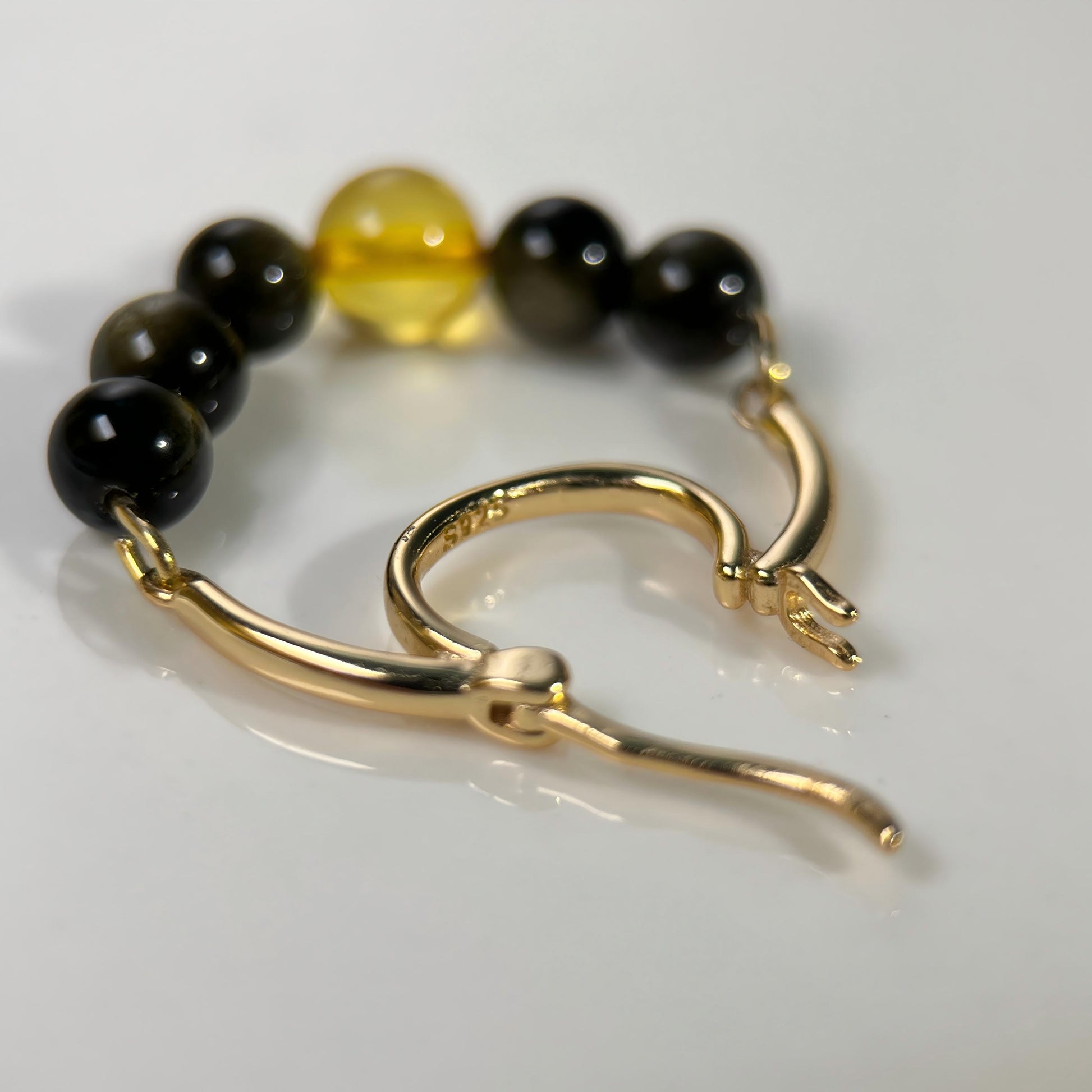 A gold-toned hoop earring with a beaded strand featuring black and translucent amber-yellow beads, photographed on a clean white surface.