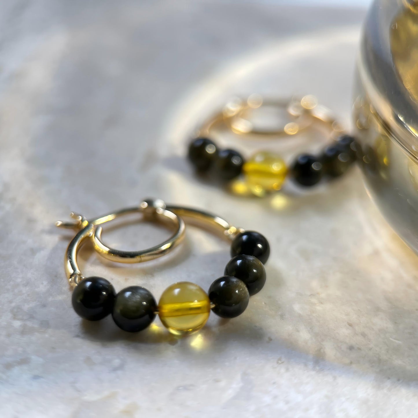Gold hoop earrings with black and translucent yellow beads, displayed on a light marble surface with a blurred gold container in the background.
