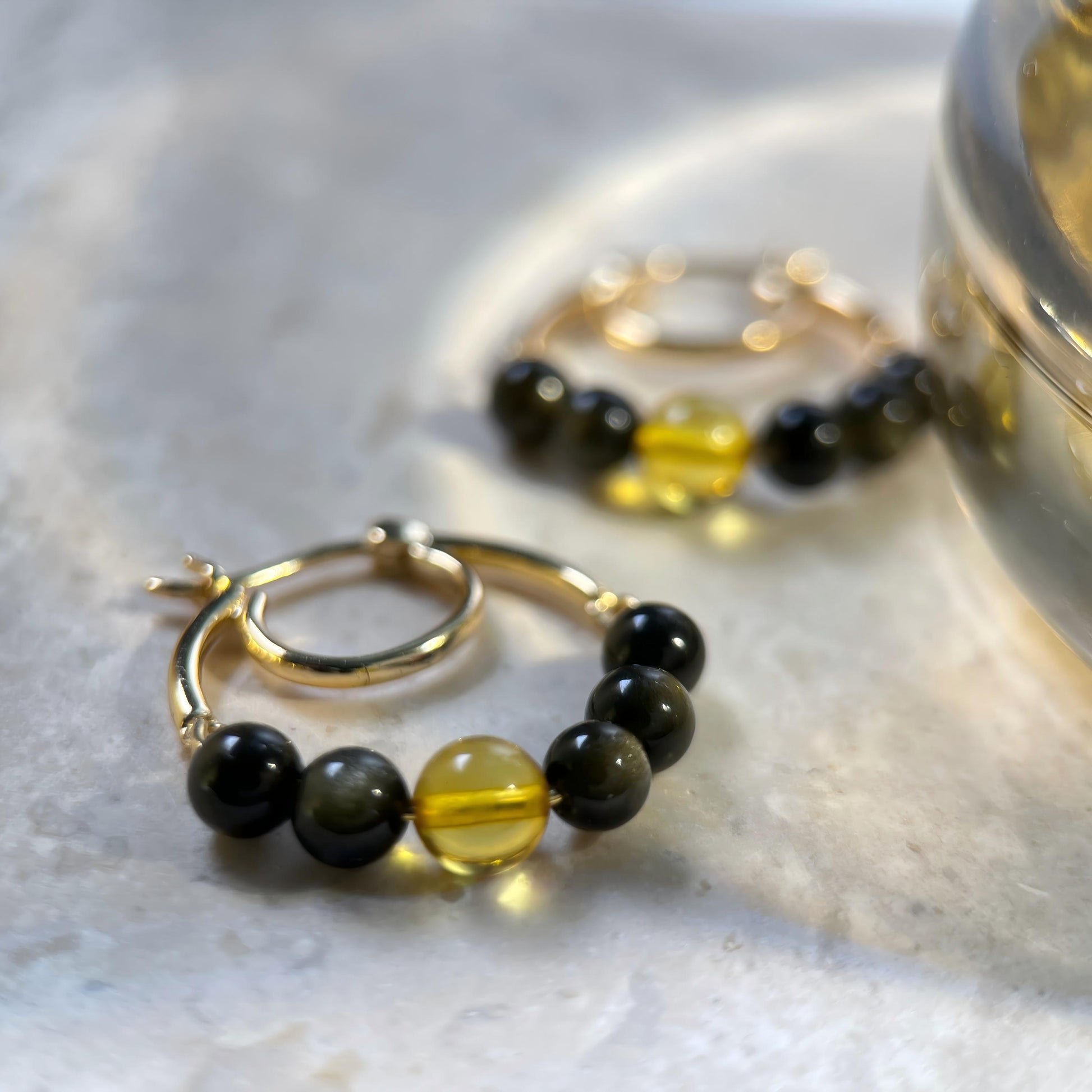Gold hoop earrings with black and translucent yellow beads, displayed on a light marble surface with a blurred gold container in the background.