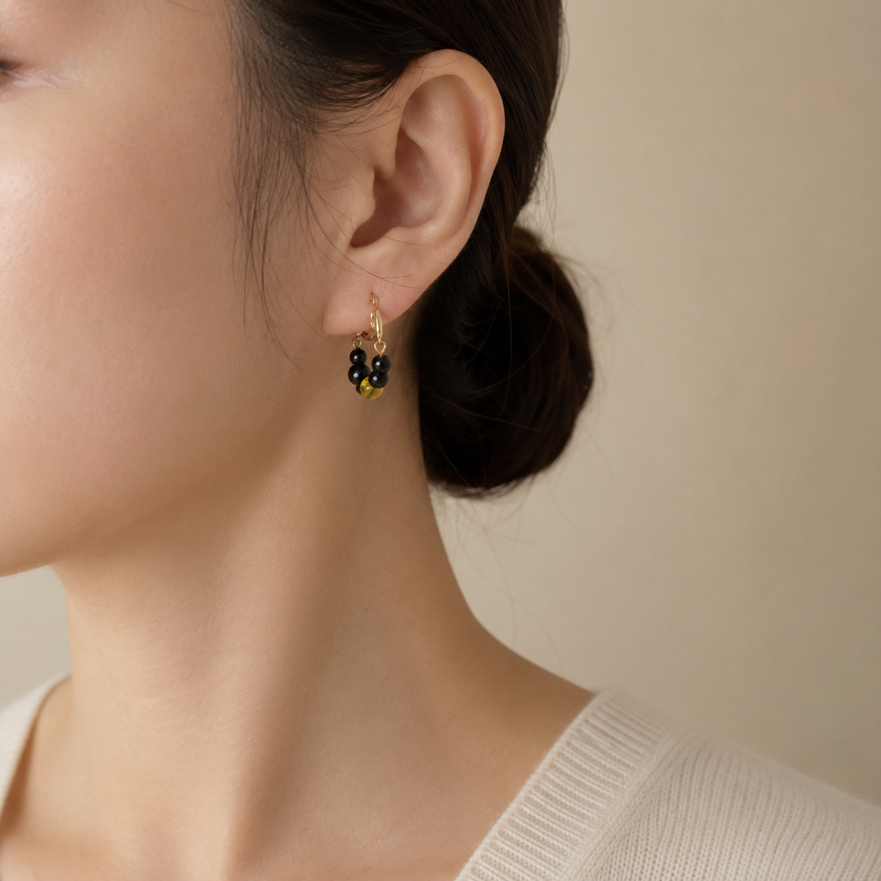 A close-up of a woman's ear wearing a small gold hoop earring with dangling black beads and tiny yellow accents, against a soft beige background.