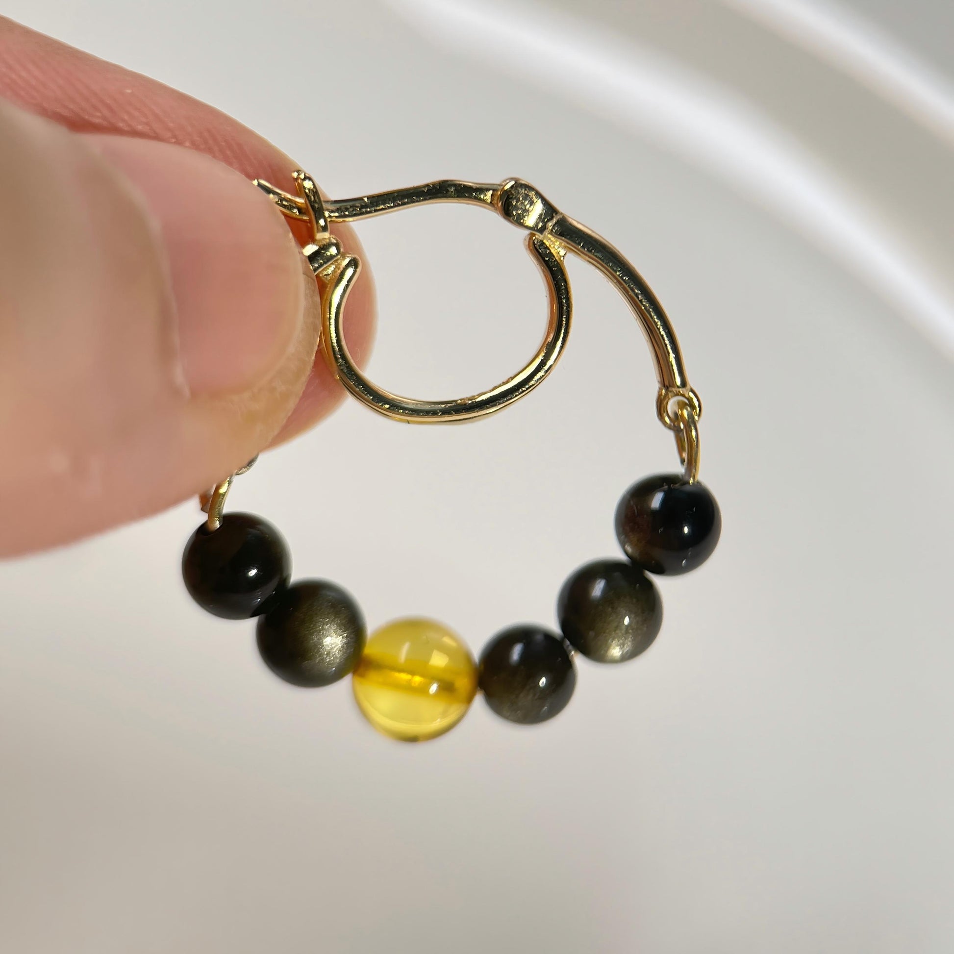 A close-up of a hand holding a small gold hoop earring with five dark round beads and one translucent yellow bead, against a soft white background.