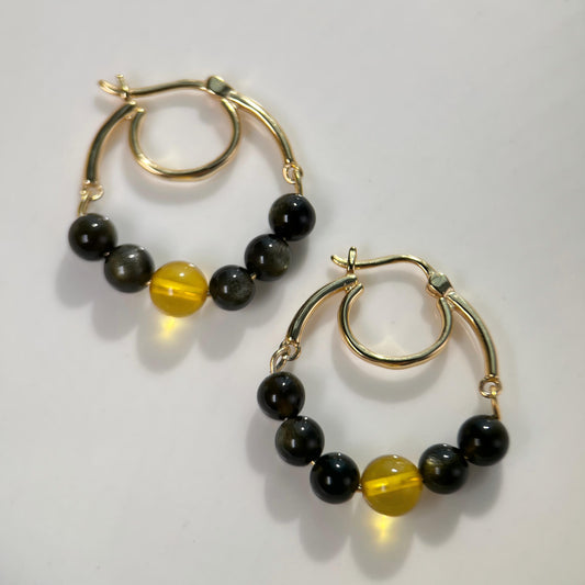 A pair of gold hoop earrings with small beaded strands featuring black beads and one translucent yellow bead, displayed on a light gray background.