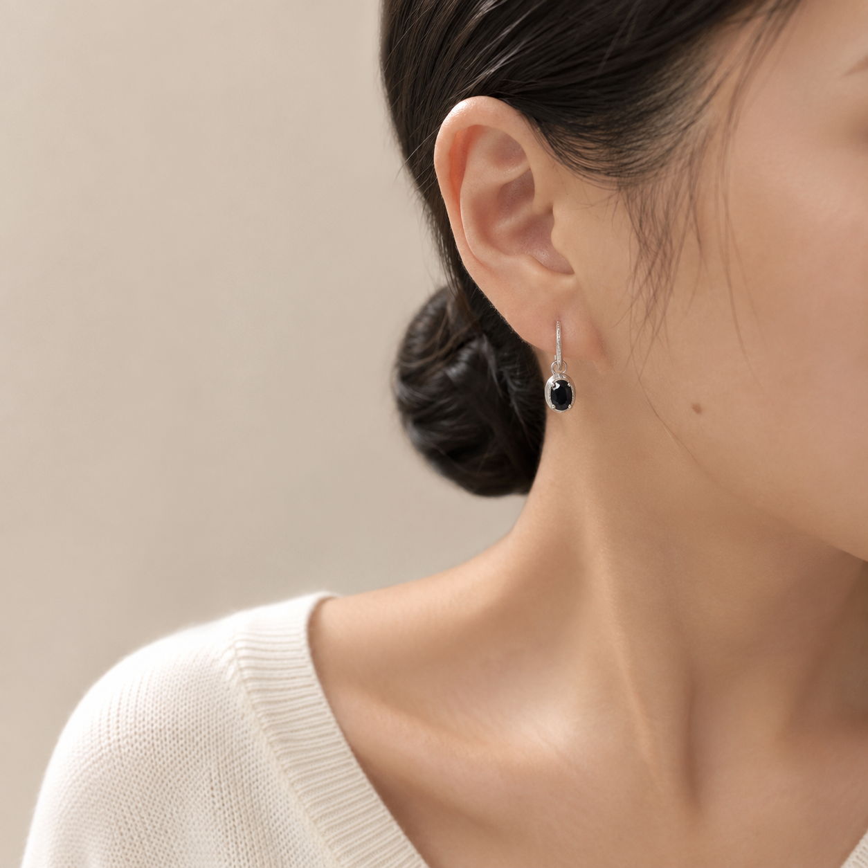 A close-up of a woman's ear wearing a delicate silver dangle earring with a black spinel stone, against a soft beige background.
